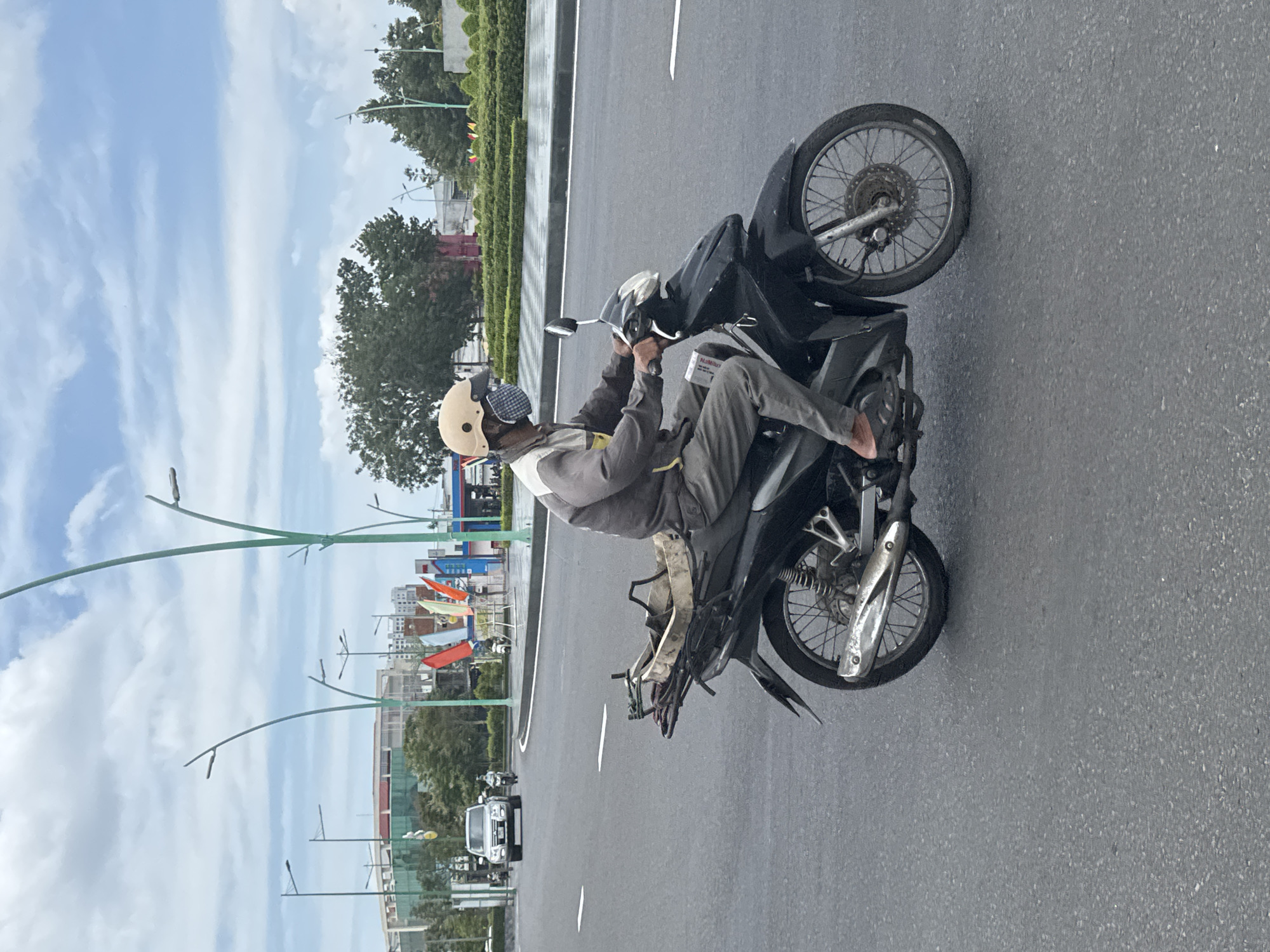 A man wearing a light tan helmet, a checkered face mask, and a gray and yellow work uniform rides a black motorcycle on a wet, asphalt road. The motorcycle has a light-colored carrier attached to the back, containing what appears to be tools and construction materials. The man is wearing dark pants and sandals, with his right foot resting on the motorcycle's footpeg. A white piece of paper is tucked into the handlebar grip. In the background, there are trees, buildings, streetlights, and flags. The sky is overcast with some patches of blue. The scene appears to be in Phan Thiết, Vietnam, a coastal city known for its beaches and historical sites.