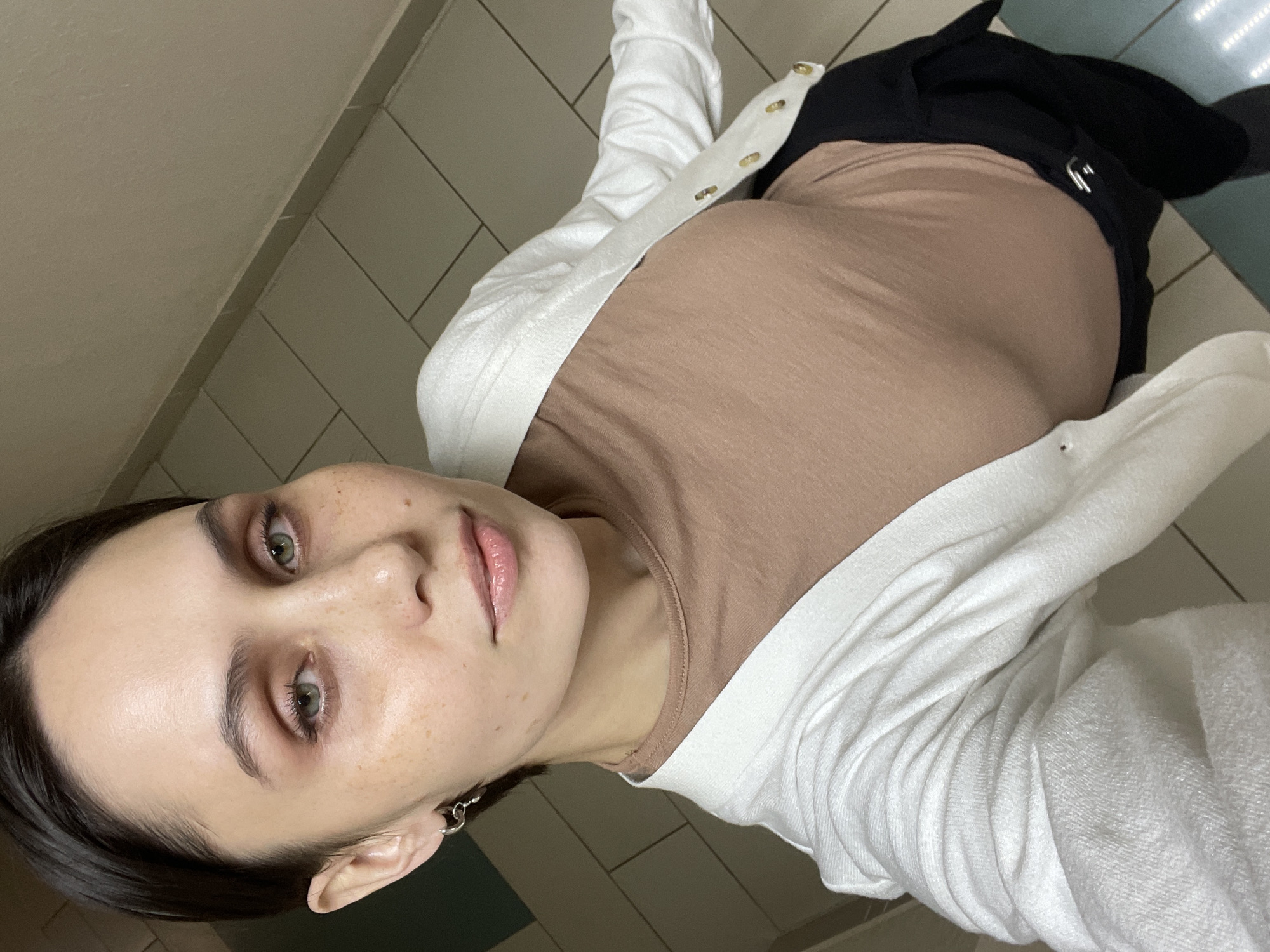 The image, captured in Izhevsk, Russia, is a self-portrait rotated 90 degrees to the left, showing a young woman looking directly at the camera. She appears to be lying down or leaning back against a wall and tiled floor. Her expression is calm and neutral, with green eyes wide open, dark eyebrows, and a subtle application of eyeshadow. She has fair skin with visible freckles across her nose and cheeks. Her dark hair is pulled back, and a small silver hoop earring is visible in her left ear, along with a delicate necklace.
She is dressed in a light brown, long-sleeved top, layered with a white knit cardigan that features several gold buttons down the front. The fabric of her top shows some gentle wrinkles. Below her waist, black trousers or a skirt are visible, paired with white socks. The background consists of light beige or gray tiled floors with dark grout lines, and a plain light-colored wall. A small section of turquoise or blue-green flooring or a mat is visible in the upper right portion of the image. The lighting appears to be soft and even, suggesting an indoor setting with artificial light. There is no visible text in the image.