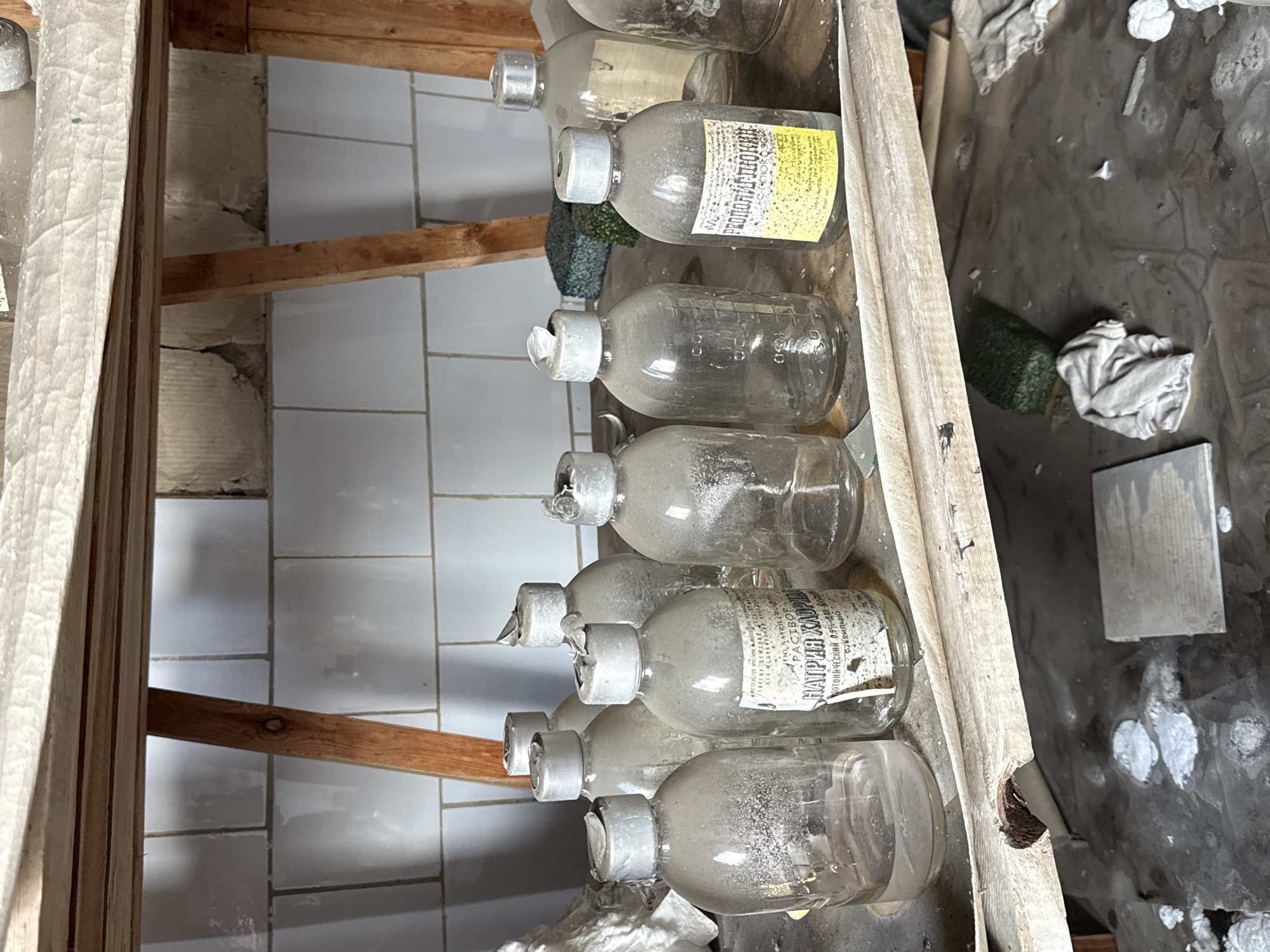 The image shows a vertical, close-up view of a shelf holding several glass bottles in an abandoned building. The bottles are dusty, some with faded labels in Cyrillic script. The setting appears to be indoors, with a tiled wall visible in the background. The overall impression is one of decay and neglect. Given the Cyrillic labels and the context of similar images, it's plausible that this scene is from an abandoned medical facility or laboratory in Prypiat, Ukraine, near the Chernobyl Exclusion Zone. The environment is derelict, with visible signs of weathering and disintegration, pointing toward long-term abandonment.