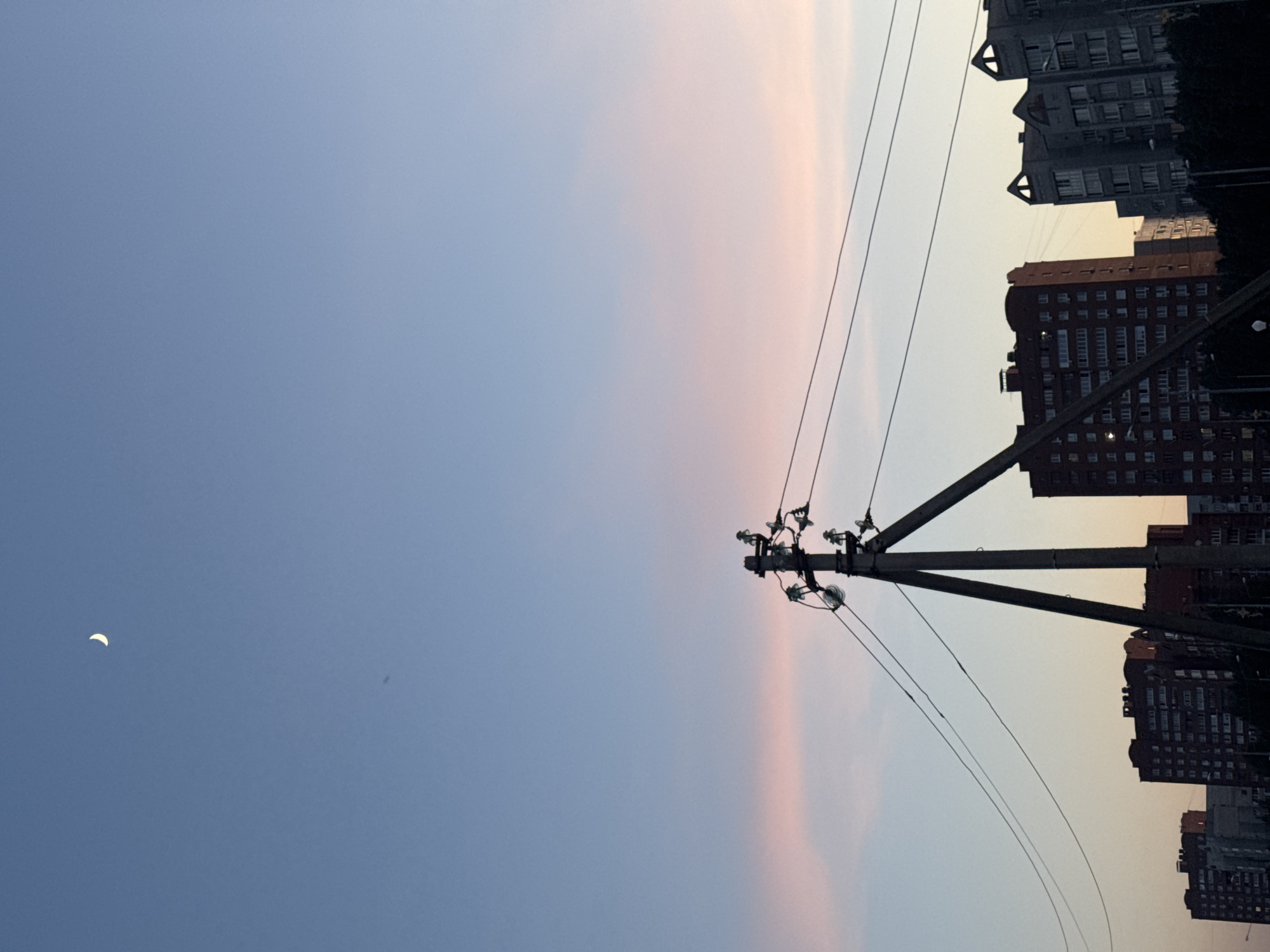 The image shows an urban scene at dusk. The sky transitions from blue on the left to shades of pink and orange towards the right. A crescent moon is visible in the blue section of the sky. In the right side of the image are buildings, silhouetted against the lighter sky, with visible windows. An electrical pole with wires is also present. The location appears to be an unidentified urban area.