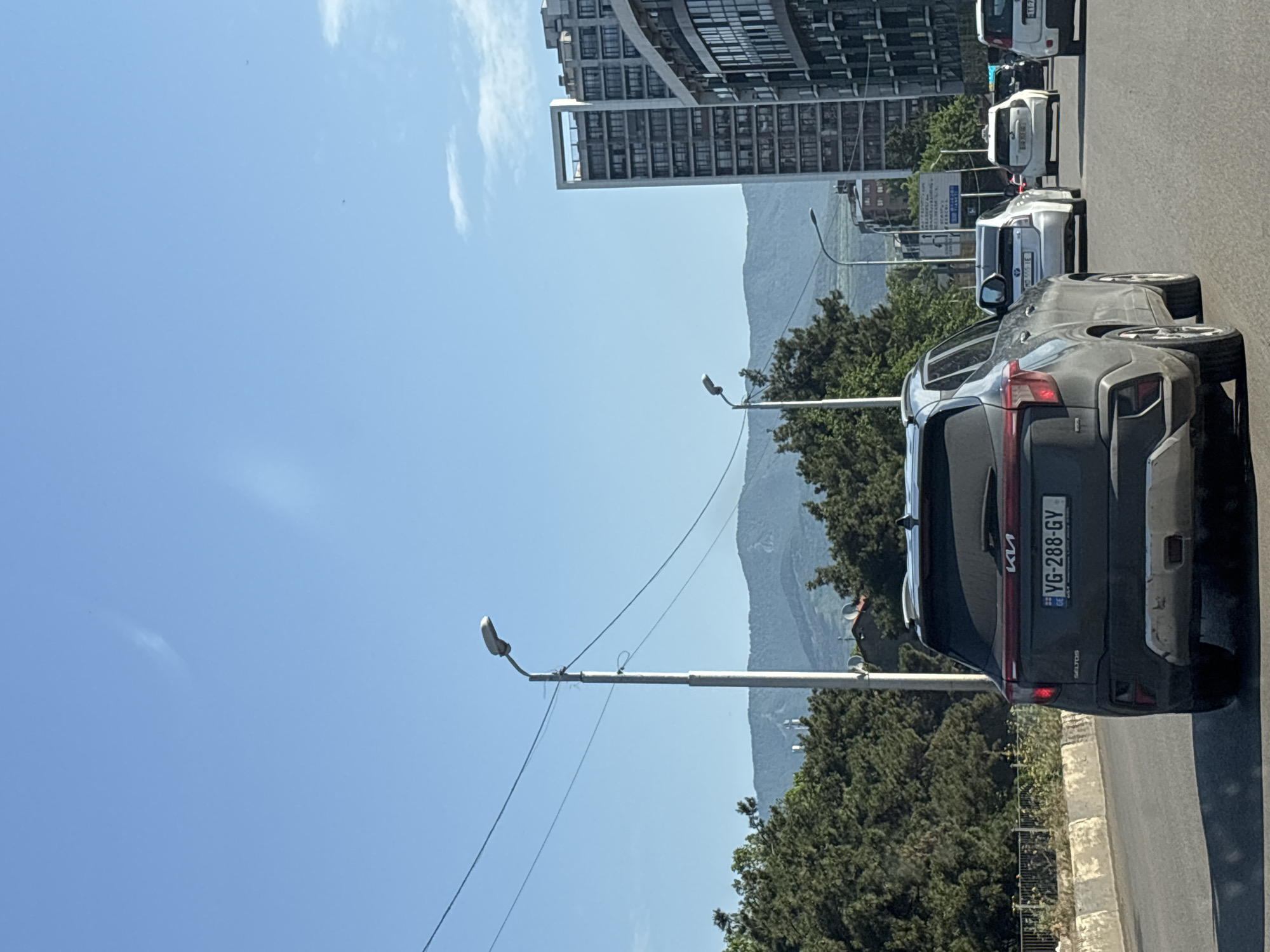 The image shows a street scene in Tbilisi, Georgia, on a clear day with a bright blue sky. In the foreground, a dark gray Kia Seltos is driving on a multi-lane road. The car's license plate reads "YG-288-GY". The road has a concrete barrier on the left. Behind the Kia, several other vehicles are visible, including white cars and a taxi. Trees and buildings line the road. In the background, distant mountains are visible, and a tall modern building stands out amongst others. A street light is positioned to the left of the road, with wires stretching across the frame. The scene appears to be a typical urban environment with traffic and infrastructure.