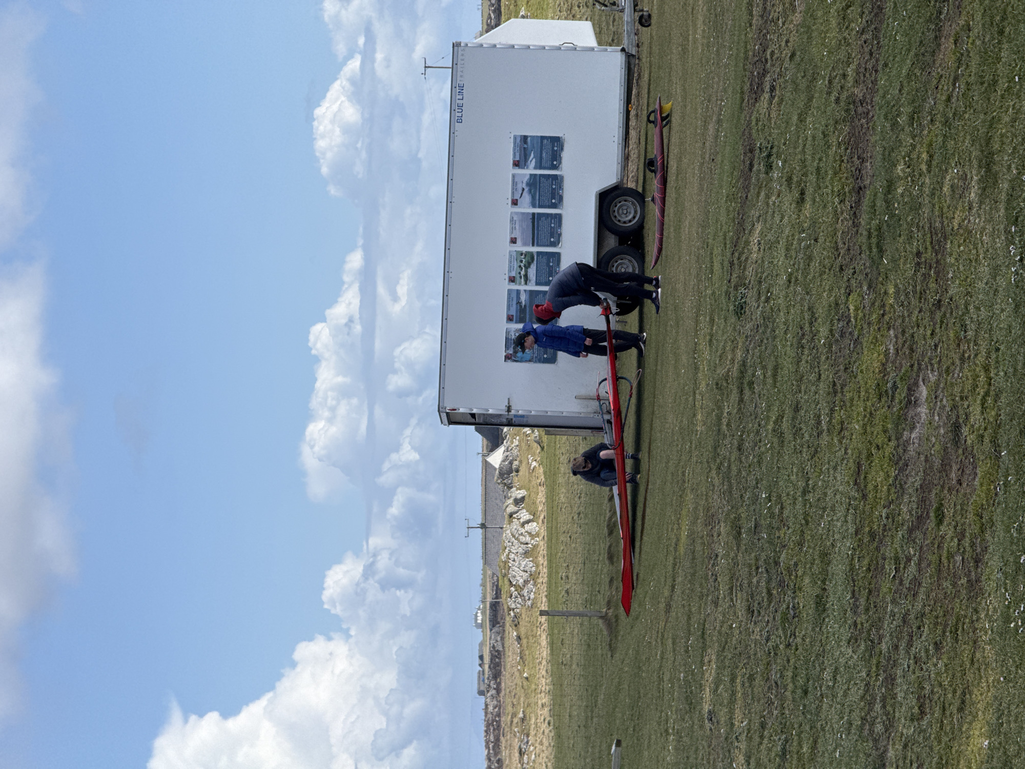 On a grassy field in Unknown, Unknown, a white trailer with "BLUE LINE" printed on the side is parked. People are interacting around a red and black windsurfing board near the trailer. Another windsurfing board is visible by the trailer's wheel. The sky is blue with cumulus clouds. In the background, a rocky terrain with some houses and a utility pole is visible.