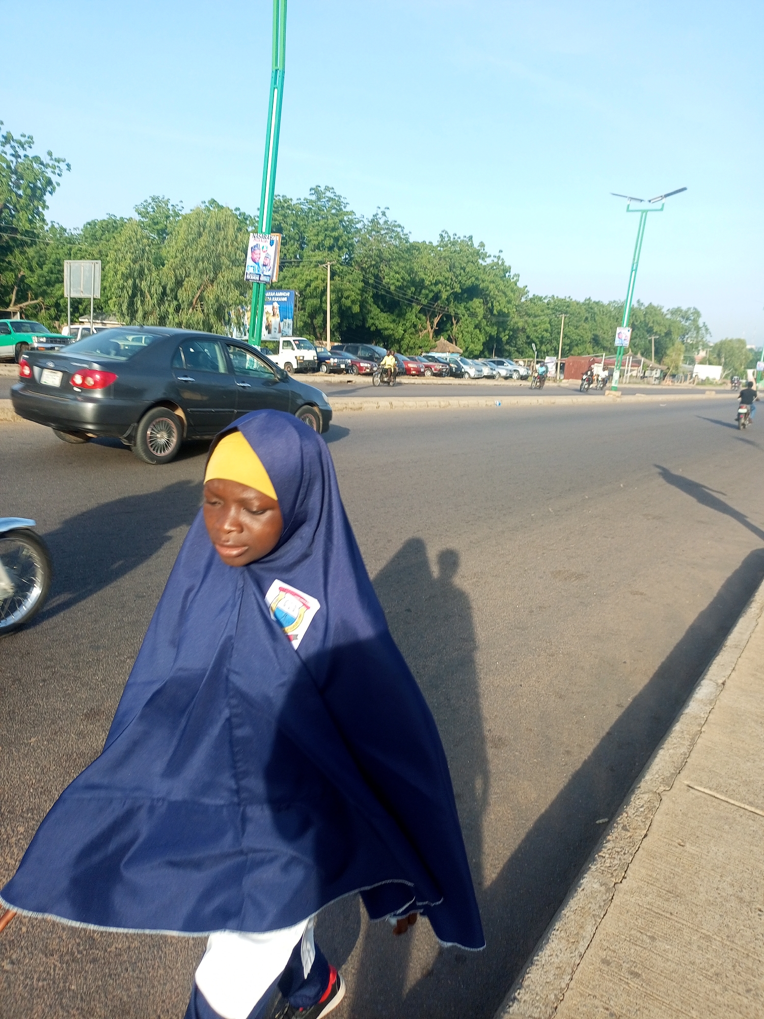 In Gombe, Nigeria, a young girl wearing a dark blue hijab and a yellow headscarf walks along a paved road under a clear blue sky. She is in the foreground, her body angled to the right, with her face downcast. A dark colored sedan is visible behind her, in the middle ground, driving away from the viewer. Several other cars and motorbikes are present in the background, along with trees and streetlights. The scene appears to be a busy street in the city.