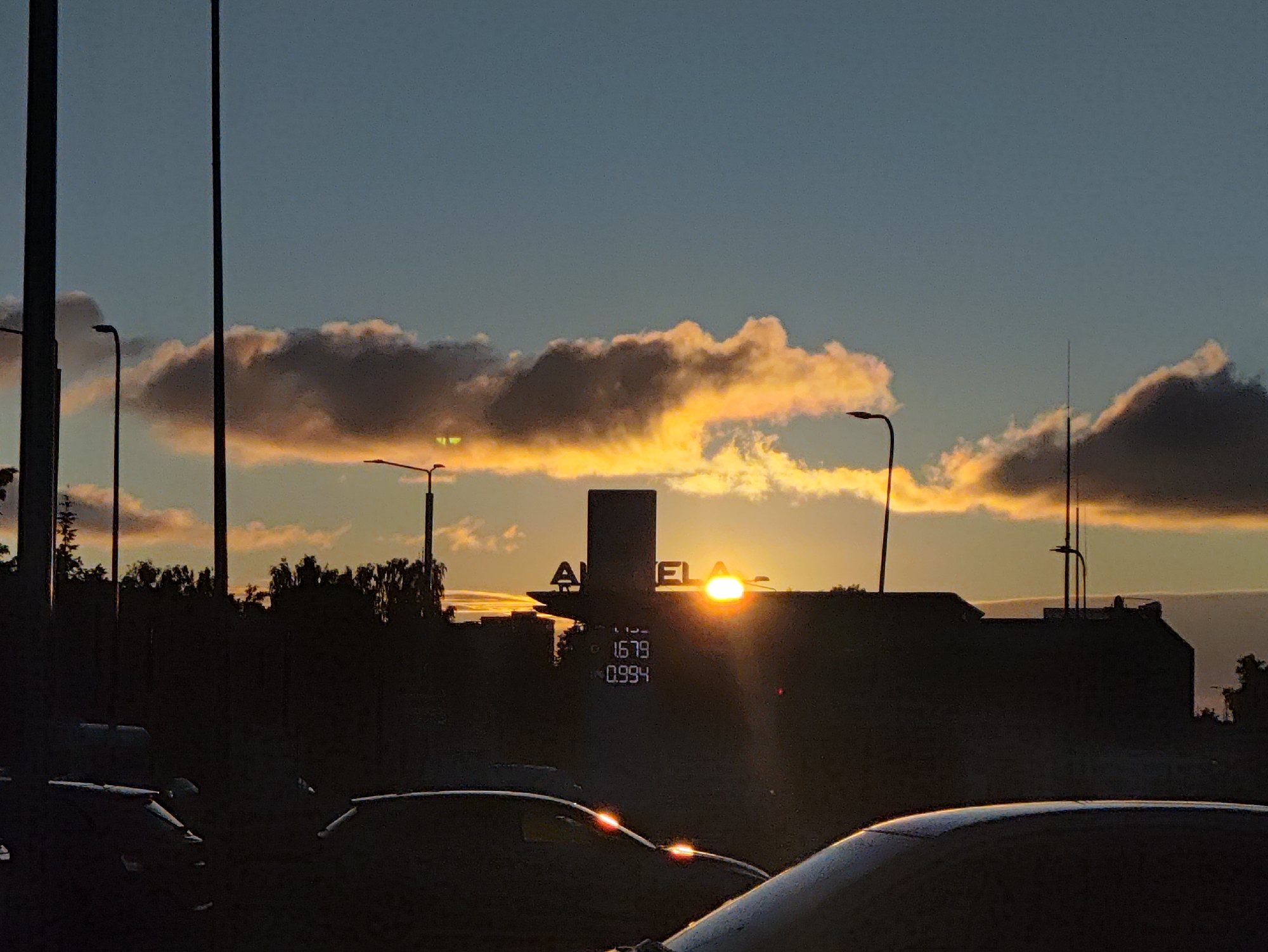 The image depicts a sunset in Tallinn, Estonia, with the sun setting behind a building. The sky is a gradient of blue and orange, with illuminated clouds. The silhouette of a building with illuminated signage is visible, showing the text "AMELA" and numerical prices "1.679" and "0.994". Several streetlights are present, and the silhouettes of trees and other buildings are also visible in the background. In the foreground, the dark silhouettes of several cars are visible. The overall scene is a twilight cityscape with a focus on the sunset and the illuminated signage.