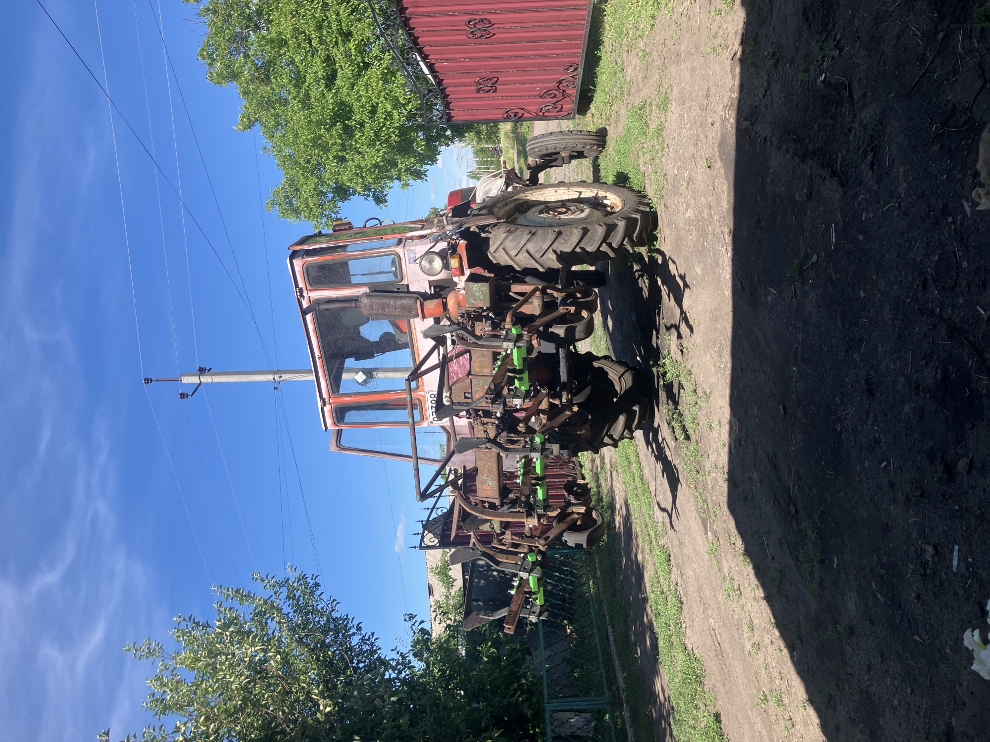 A red and white tractor with agricultural implements attached is parked on a dirt road in Vovkivka, Ukraine. The tractor is in the foreground, facing the viewer, and is positioned between two red metal gates. The gates are ornately decorated and appear to be part of a fence. To the left of the tractor, there is a green fence and lush green trees. The sky is clear and blue with some wispy white clouds. A power pole with overhead wires is visible behind the tractor. The overall scene suggests a rural setting, likely a farm or a residential property with agricultural equipment. The tractor appears to be weathered and possibly old, but functional.