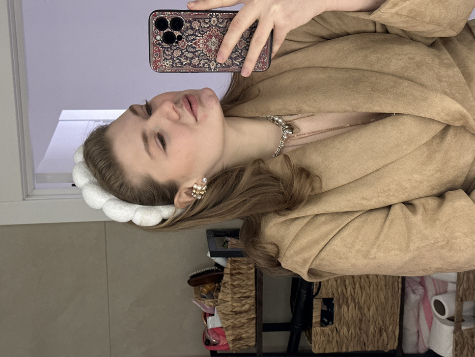 A young adult female is depicted in a mirror selfie, positioned indoors in what appears to be a bathroom in Kazan, Russia. The subject has light brown hair, pulled back by a prominent white padded headband. She wears a large, multi-stone ear cuff featuring pearl-like and clear elements on her visible right ear, and a gold-toned necklace with a silver-colored chain and a heart-shaped pendant. She is dressed in a light brown, possibly faux-suede, open-front jacket or robe. Her right hand, partially visible, holds a smartphone with a dark, ornate patterned case resembling a Persian rug. She is looking towards her reflection in the mirror, with her lips slightly parted. The background reveals a light-colored wall with a horizontal seam, a partially open white door frame, and a shelving unit. On the shelves, two woven wicker baskets contain various items, including personal care products and possibly hairbrushes. Below the baskets, two white rolled towels with pink stripes and two rolls of white toilet paper are visible.