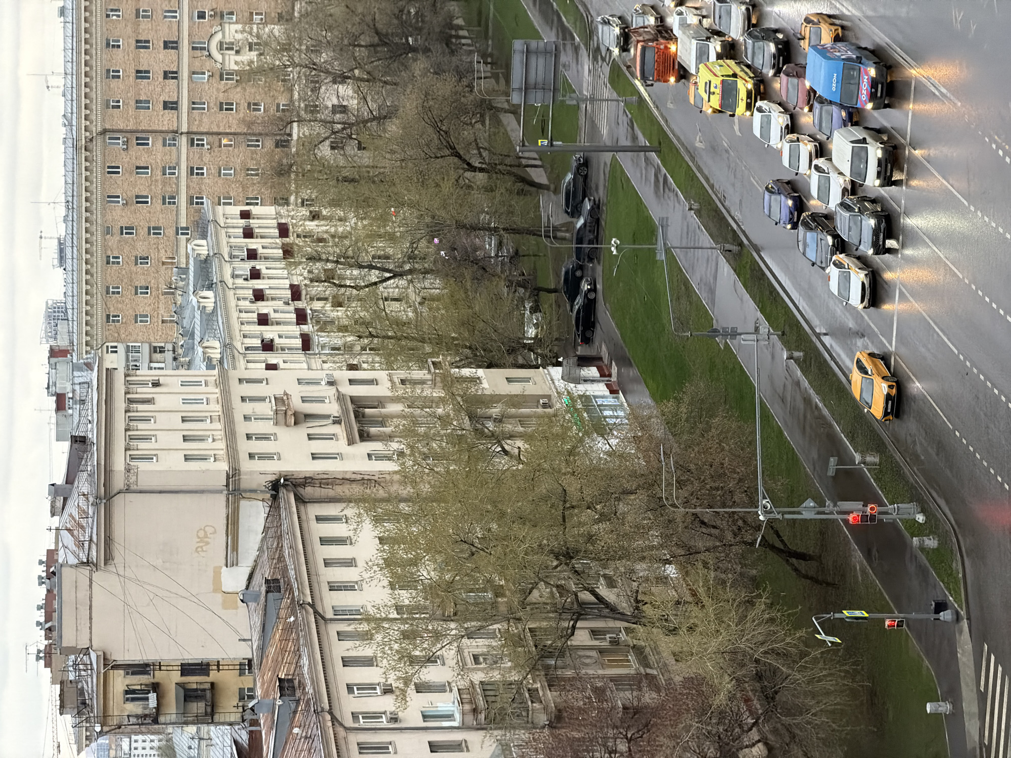 The image presents an elevated view of a street in Moscow, Russia. Tall buildings line the left side of the frame, while the street, filled with vehicles, dominates the right side. The vehicles include vans, cars and at least one yellow taxi. A traffic light displays a red signal. Between the street and the buildings is a strip of greenery with trees. The scene is overcast with likely wet conditions.