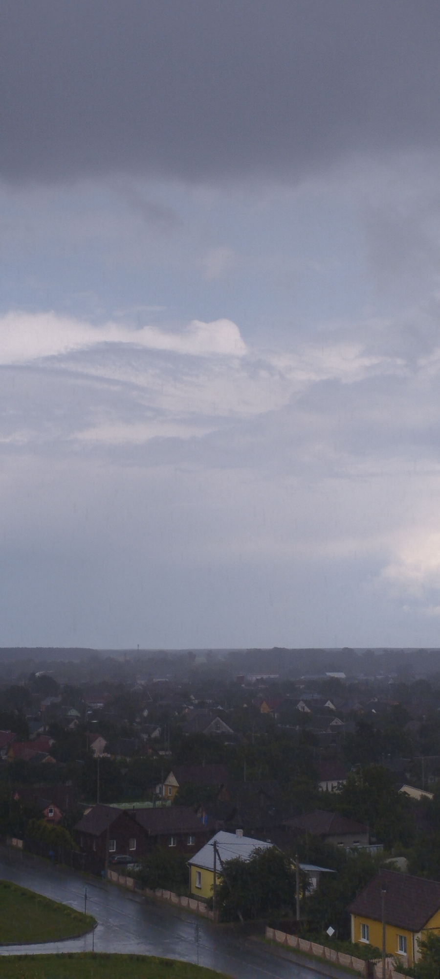 This is a full-frame, high-angle, vertical, outdoor, daytime shot of a residential area in the city of Skidzieĺ, Belarus, under overcast and possibly rainy conditions. The sky occupies the upper half of the image, filled with dark, heavy clouds at the top and lighter, more diffused clouds lower down, with visible rain streaks. The lower half of the image depicts a dense collection of houses with dark roofs, interspersed with trees and greenery. A paved road, wet from rain, is visible in the foreground, curving to the left. Several houses are distinct, including a dark-roofed house with white-framed windows and doors, a bright yellow house with a light-colored roof and visible windows, and another yellow house with a brown roof further to the right. The overall impression is of a somber, somewhat dreary atmosphere characteristic of inclement weather in a suburban or rural residential setting.
