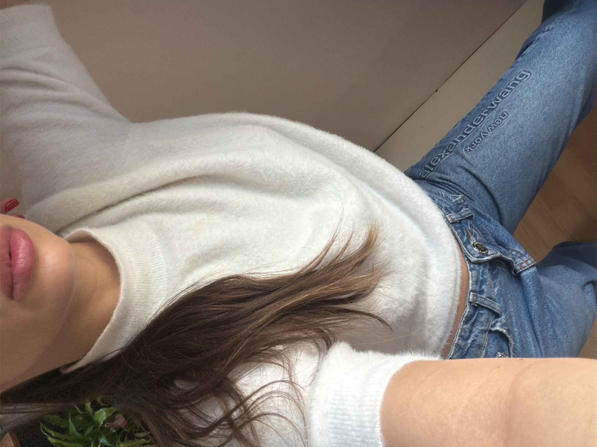 An indoor, chest-down portrait shot captures a female individual in Sofia, Bulgaria; no specific landmark or street is identifiable from the visible details. The subject's head is partially cropped, revealing the lower face including the chin, mouth with pink-tinted lips, and neck. Long, brown hair with lighter highlights cascades over her left shoulder and against a white, fuzzy, long-sleeved mock-neck sweater. The left forearm is visible, angled upward as if holding a camera. She is wearing blue denim jeans, on the right leg of which "alexanderwang" and "New York" are debossed or embroidered vertically. The background on the left features a portion of a green potted plant with small pinkish elements. To the right, a plain light brown wall is visible, with a section of a light wooden floor at the bottom right. The scene is brightly and evenly lit.