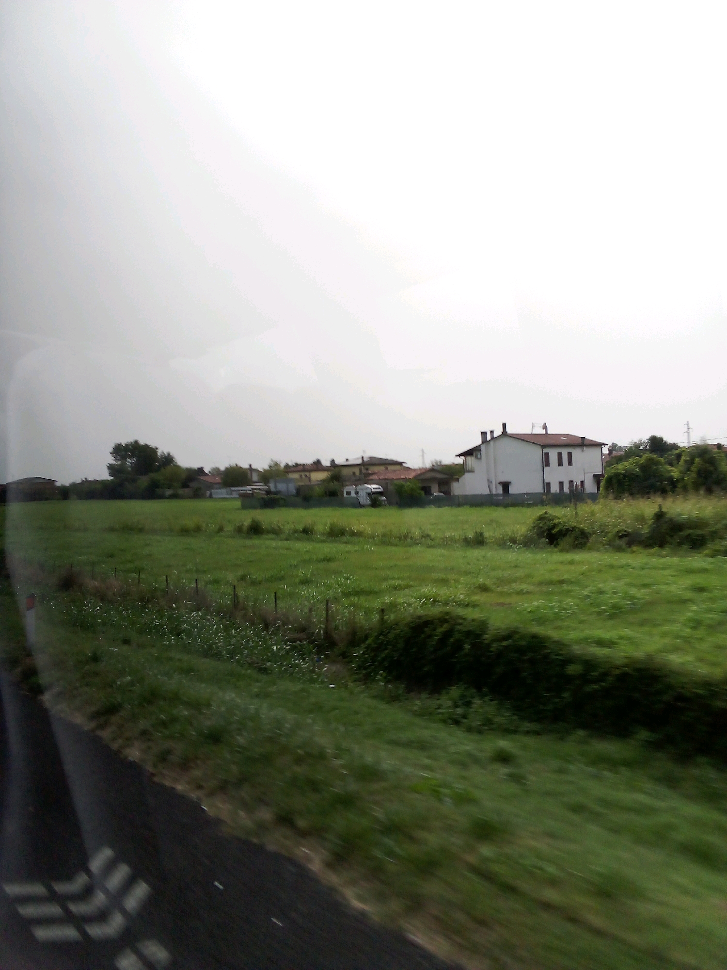 This is a medium-shot, eye-level, outdoor, daytime, slightly blurry photograph taken from a moving vehicle. The scene captures a rural landscape near Solesino, Italy. A white, two-story house with a red tile roof and dark windows is the dominant structure, situated in the middle ground on the right. It has several chimneys. To the left of this house, there are other residential buildings with red roofs and a white recreational vehicle or camper van. A large, green field stretches across the foreground and middle ground, with some scattered wild flowers and a low fence line. In the far left of the image, a dark asphalt road with white lane markings is visible. The sky is overcast and bright. The overall impression is one of a tranquil, agricultural area.