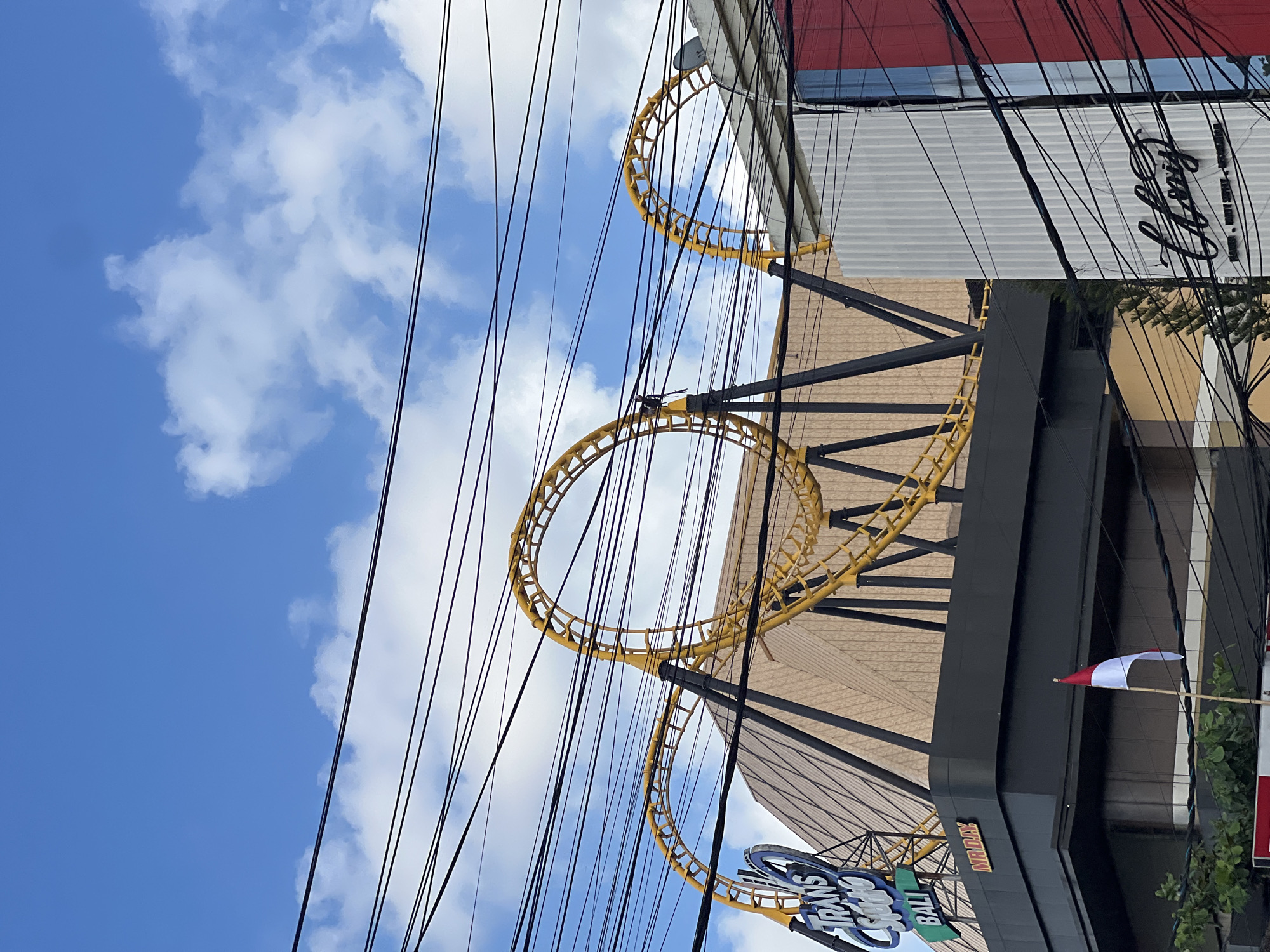 The image depicts a street-level view of the Trans Studio Bali amusement park, a prominent attraction in the city of Denpasar, Indonesia. The foreground is dominated by a dense network of black overhead electrical wires, crisscrossing the frame and partially obscuring the view of the structures behind them.
Dominating the mid-ground is a yellow roller coaster track, featuring loops and curves, supported by black metal framework. This roller coaster is situated in front of a large, pyramid-shaped building, likely part of the amusement park's main structure. To the left, a blue sign with white lettering reads "TRANS STUDIO BALI," identifying the location. Below this sign, on a black structure, is a smaller sign with red lettering that says "MR.DIY."
To the right of the roller coaster, a white building with vertical corrugated panels is visible. A section of a red building is also present on the far right edge of the image. In front of the black structure and below the "TRANS STUDIO BALI" sign, a small Indonesian flag, red and white, is mounted on a bamboo pole, attached to a white object. Lush green foliage is visible in the lower left corner, suggesting the presence of trees. The sky above is a clear blue, interspersed with scattered white clouds, indicating daytime.