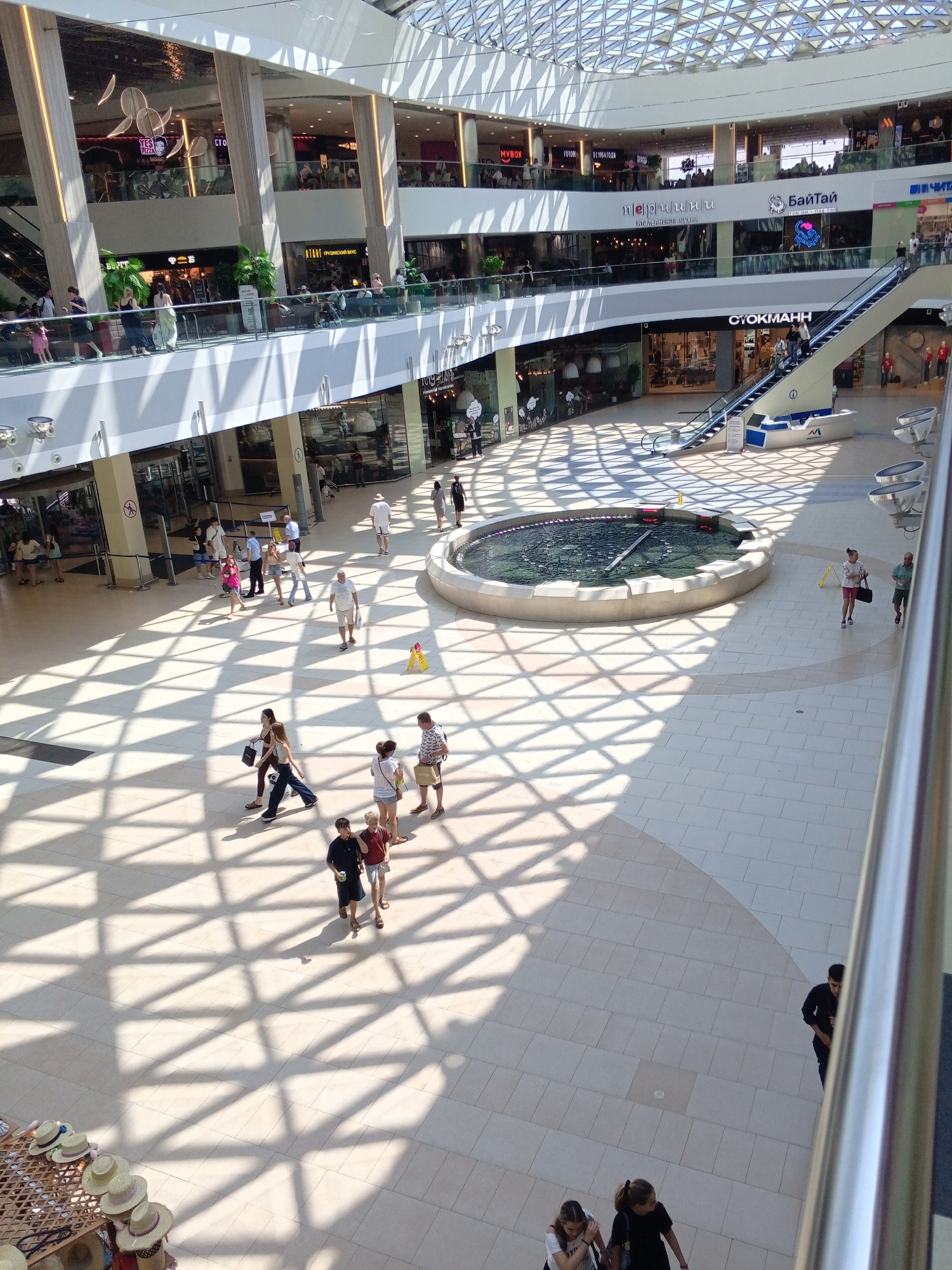 The image depicts the interior of a modern, multi-level shopping mall, likely in Sochi, Russia, characterized by its spacious atrium and large, geometric skylight that casts a diamond-patterned shadow across the tiled floor.
In the foreground, several individuals are walking across the open floor. Two young women in black and white tops are walking away from the camera, one carrying a black bag. Further back, a young man in a black t-shirt and dark shorts walks with another young person wearing a red top and shorts. A few other people are visible in the mid-ground, including a man in a white t-shirt and shorts walking towards a circular fountain, and a woman in a pink top and white shorts walking near the fountain with a black bag. There are also groups of people in the upper levels of the mall, visible through storefronts and on walkways.
The environment includes a large, circular fountain with a dark water surface in the center of the atrium. Escalators are visible on the right side of the frame, and various storefronts with illuminated signs are located on multiple levels. To the left, a display of straw hats on stands is visible at the bottom of the image. The overall impression is of a busy, well-lit commercial space.