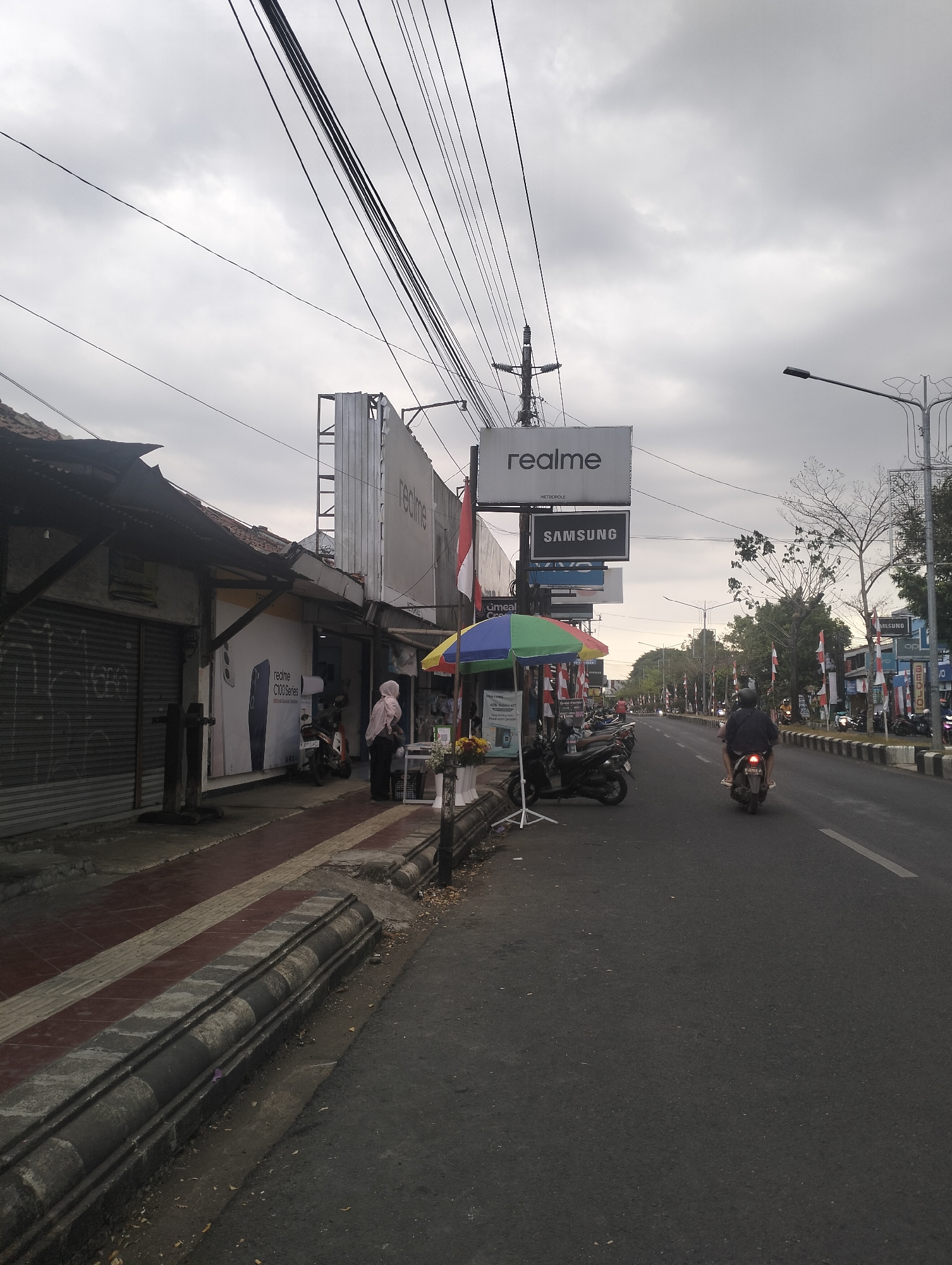 A street scene in Kandang Gampang, Indonesia, shows a cloudy sky above a row of buildings on the left and a road on the right. The buildings appear to be shops, with several prominent signs visible, including "realme" and "SAMSUNG". A woman in a pink hijab stands on the sidewalk in front of one of the shops, next to a display of umbrellas and flowers. Several motorcycles are parked on the sidewalk and a few are on the road. One motorcycle with a rider is moving away from the camera down the street. The road has a white line marking its edge and a motorbike lane. The overall impression is of a commercial street on an overcast day.