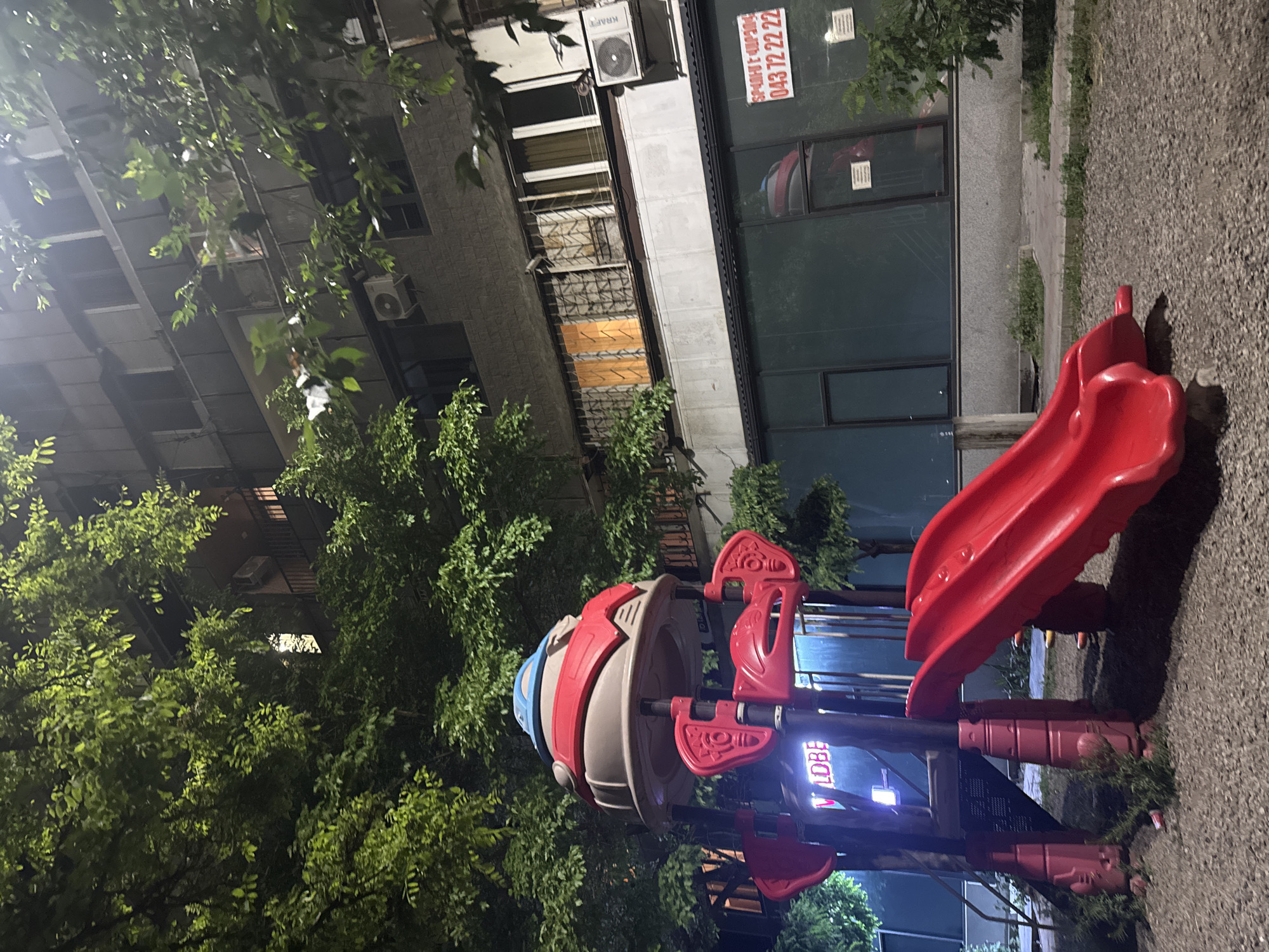 The image depicts a playground structure with a red slide and a spinning carousel-like apparatus, located outdoors at night in Yerevan, Armenia. The playground equipment is adjacent to a building with multiple floors, some windows having what appear to be security bars. A sign with the numbers "043 72 22 22" is visible on the building's exterior. Lush green foliage partially obscures the building and playground equipment, indicating the presence of trees. The ground is covered with a rough, gravelly surface, typical for playgrounds. There are no people visible in the frame. The light suggests the playground is artificially lit, with some illumination coming from the building.