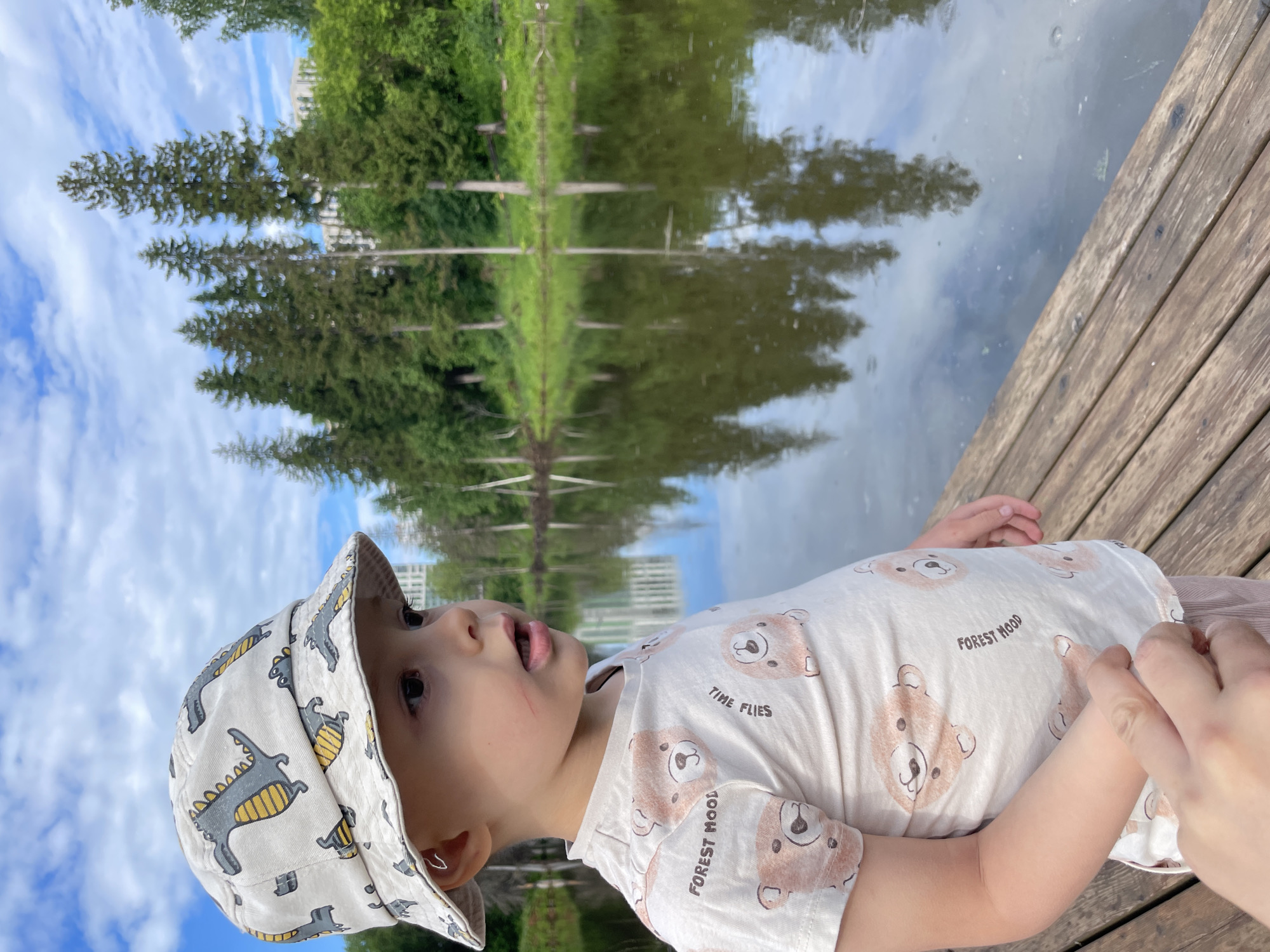 A young child wearing a bucket hat with a dinosaur pattern and a t-shirt with teddy bear prints stands on a wooden pier next to a lake. The child is looking to the right, with their mouth slightly open. The lake is surrounded by trees and greenery, with buildings visible in the distance. The sky is blue with white clouds, and the trees are reflected in the calm water of the lake. The overall scene is one of a peaceful outdoor setting. There is no specific information in the image to indicate the location is in Izhevsk, Russia.