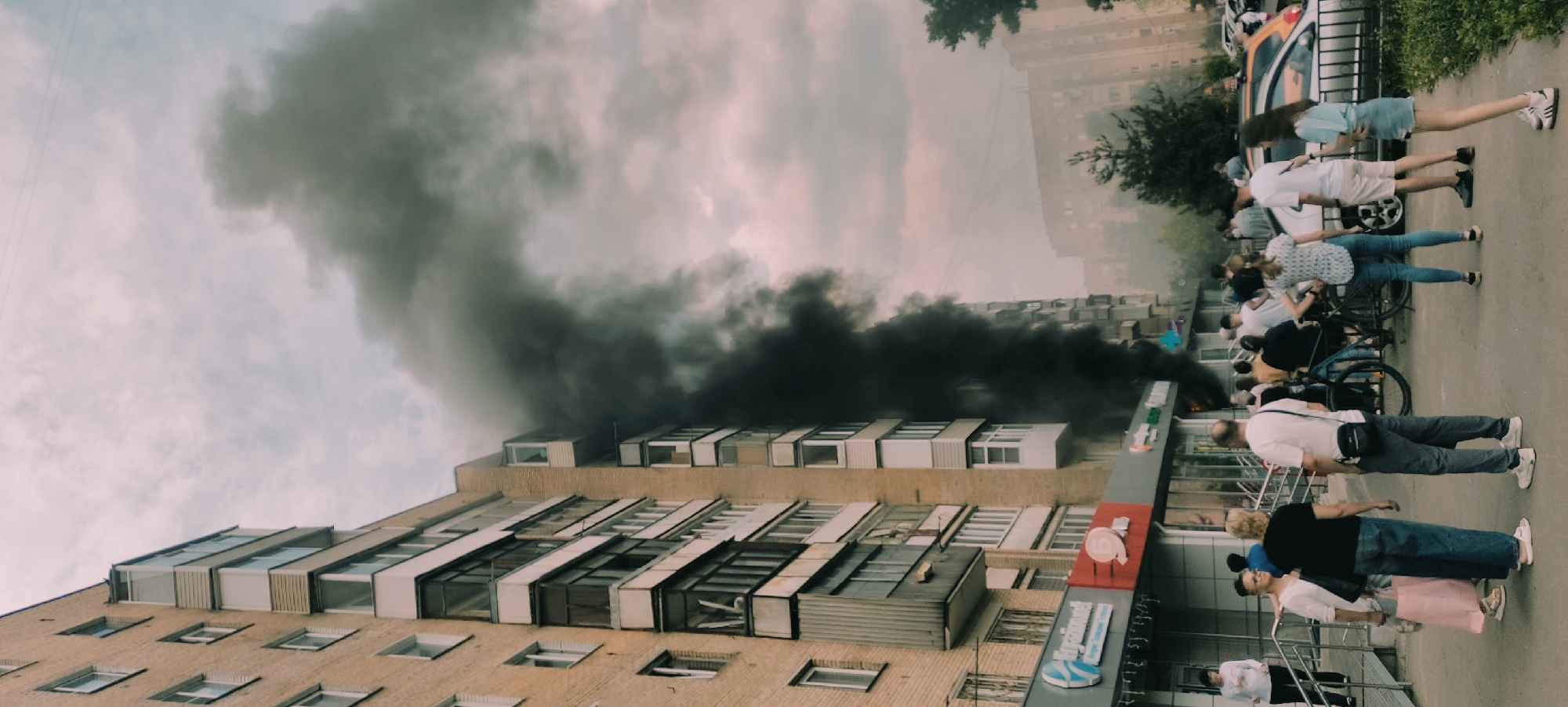 In an urban area, a building is emitting a significant plume of black smoke. Several people are standing on the street, observing the scene. The building appears to be multi-storied and of brick construction. The environment suggests a fire or other incident causing the smoke.