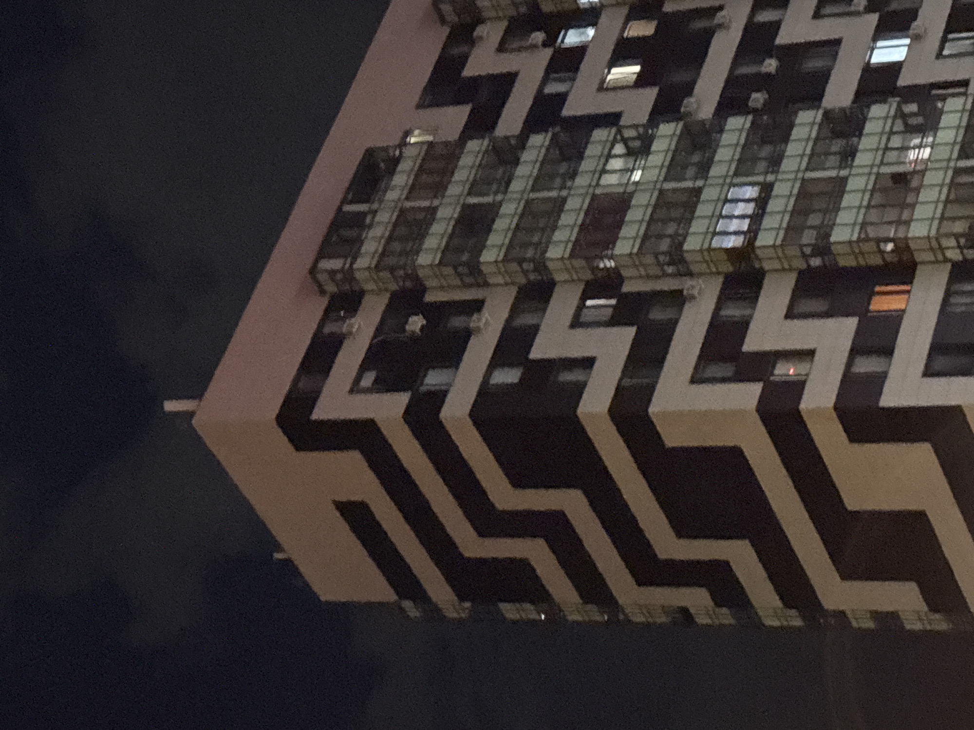 This is a nighttime, low-angle, close-up shot of a modern apartment building in Kazan, Russia. The building features a striking geometric facade with alternating dark brown and beige zig-zagging bands. A section of the building is characterized by a glass-fronted tower with multiple floors of windows and balconies. Some windows are illuminated, indicating occupancy. The sky above is dark and cloudy.
