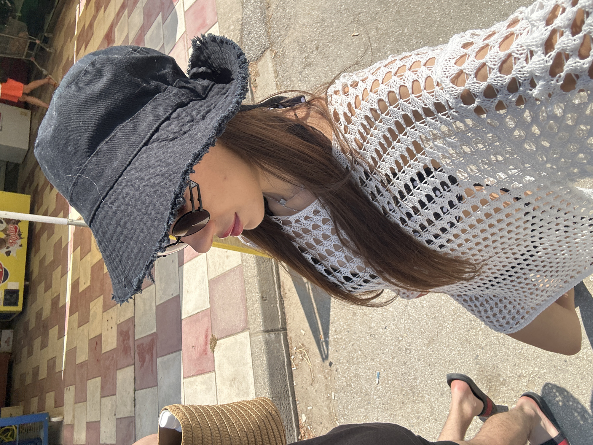 In Ofrynio Beach, Greece, a young woman wearing a black bucket hat, sunglasses, and a white crochet top walks outdoors on a sunny day. She is seen from the chest up, her long brown hair flowing. To her left, another person's legs are visible, wearing black flip-flops. The background features a tiled walkway, some vegetation, and a yellow kiosk.