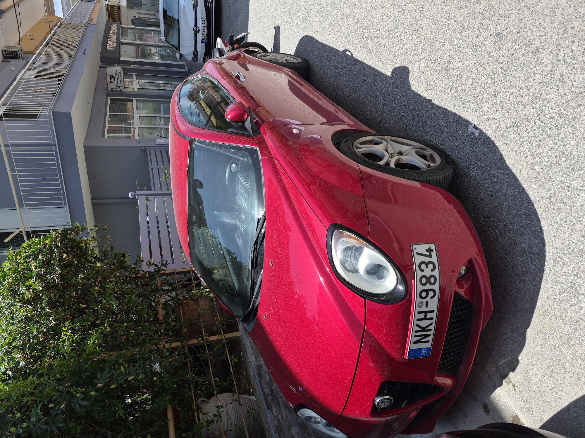 A red Alfa Romeo MiTo is parked on a paved street in front of a two-story building in Νέα Καλλικράτεια, Greece. The car's license plate reads "NKH-9834". The building has a gray facade, a white balcony railing on the upper floor, and shops on the ground level, one of which has a sign that reads "Μαγαζί 24ωρο Τεμπονου". A white Volkswagen Golf is parked behind the Alfa Romeo. Green foliage and trees are visible behind a yellow fence to the left of the Alfa Romeo. Sunlight casts strong shadows on the ground.