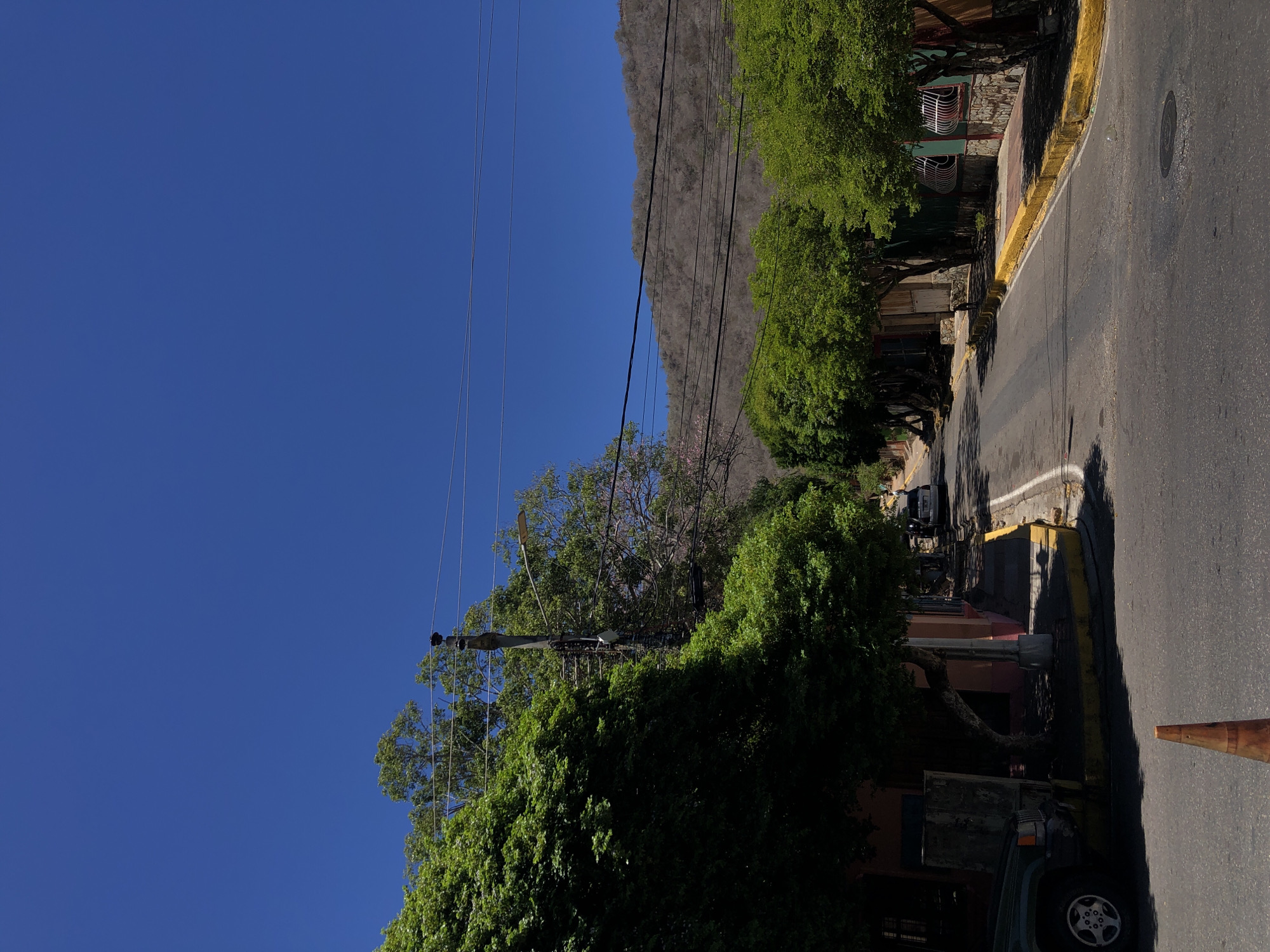 The image captures a sunlit street scene, likely set in El Valle del Espíritu Santo, Venezuela, showcasing a quiet residential or semi-residential area against a dramatic mountain backdrop.
The upper left portion of the frame is dominated by a clear, deep blue sky, indicating bright, fair weather. Stretching across the background and mid-left, a large, rocky mountain range, appearing somewhat arid with sparse vegetation, provides a imposing natural boundary to the scene.
Running along the right side of the image is an asphalt road, bordered by a concrete sidewalk with a vibrant yellow-painted curb. A circular manhole cover and a faint white line are visible on the road surface. In the lower right, a dark-colored SUV or jeep is parked, with its front wheel and bumper partially in view, next to a rusty orange traffic cone lying on its side. Further down the street, another dark vehicle is partially obscured by dense foliage.
Numerous lush green trees line both sides of the street, with a particularly dense cluster on the far left. These trees cast long, distinct shadows across the road and sidewalks, indicative of strong sunlight, suggesting the time of day is likely late morning or early afternoon. Behind the trees on the right, several buildings or houses are visible, featuring varied architecture, including some with stone elements and windows fitted with protective wrought-iron bars. A network of utility poles and multiple power lines traverses the sky and trees, extending across the scene.
The scene is tranquil, with no people or specific activities taking place. No discernible text is visible within the image. The combination of the clear skies, mountain, and the specific urban and natural elements strongly cues to its Latin American location.