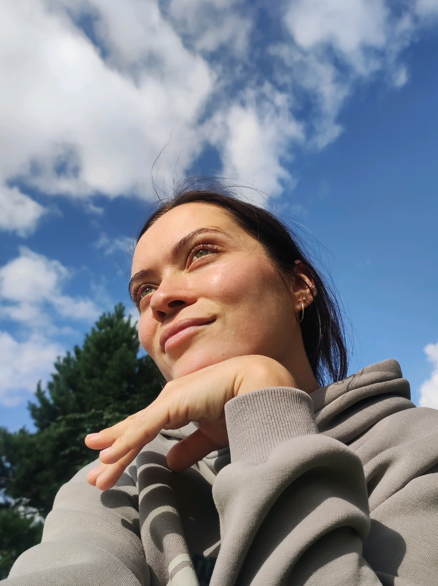 The image shows a close-up, low-angle shot of a young woman looking up and to her right, with her chin resting on her hand. She is wearing a grey hooded sweatshirt. Her dark hair is pulled back, and she has light-colored eyes. The background consists of a bright blue sky with scattered white clouds and the top of a dark green coniferous tree on the left side. The lighting suggests it is daytime, possibly late morning or early afternoon, with the sun illuminating the woman's face from the side and slightly from above. There is no indication of the specific location within the city of Фоки, Russia.