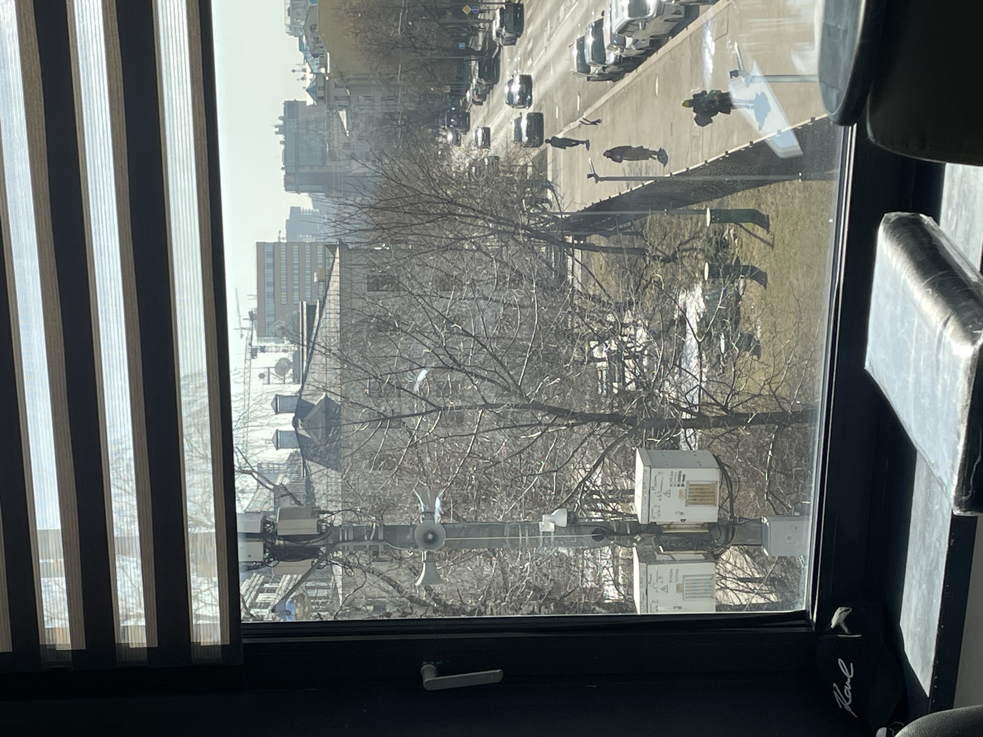 The image depicts an exterior urban scene viewed from a window, positioned on Bolshaya Pirogovskaya Street in Moscow, Russia.
The internal foreground shows a dark window frame. A gray window handle is on the left frame. A black baseball cap with "Karl" written in white script rests on the dark windowsill. A dark, possibly leather, object is partially visible on the bottom right. Above the outdoor view, a horizontally striped window blind, featuring alternating opaque dark and translucent light sections, is partially lowered. Reflections of interior elements are visible on the lower glass.
The midground outside reveals a wide multi-lane asphalt street, Bolshaya Pirogovskaya, with vehicular traffic moving in both directions. Numerous passenger cars, primarily sedans and SUVs, are present on the road and parked parallel to the curb on the right side. Several pedestrians are walking on the sidewalk adjacent to the parked cars. One person in a dark coat walks away from the viewer, and another in a lighter coat walks towards the viewer. A third person, wearing a bright yellow/green hat and carrying a bag, walks away, with their reflection visible on the window glass.
To the left of the street, a raised park area is separated by a stone retaining wall. This area contains numerous deciduous trees with bare branches, suggesting late winter or early spring. Patches of snow are visible on the grass within the park. A prominent gray utility pole, positioned in the left midground, supports multiple devices: two large, conical loudspeakers pointing horizontally, a smaller surveillance camera below them, and several white electrical utility boxes (two prominent ones are labeled "Теле 2" and "Москва"). Various wires are attached to the pole.
The background features a diverse urban landscape under a hazy bright sky. To the left, a multi-story building with a light-colored facade and a dark roof adorned with chimneys is visible behind the bare trees. Further in the distance, a mix of residential and commercial buildings of varying heights and architectural styles line the street. Construction cranes are discernible on the distant horizon, indicating ongoing development. Sunlight is evident, casting shadows and creating reflections on vehicle surfaces.