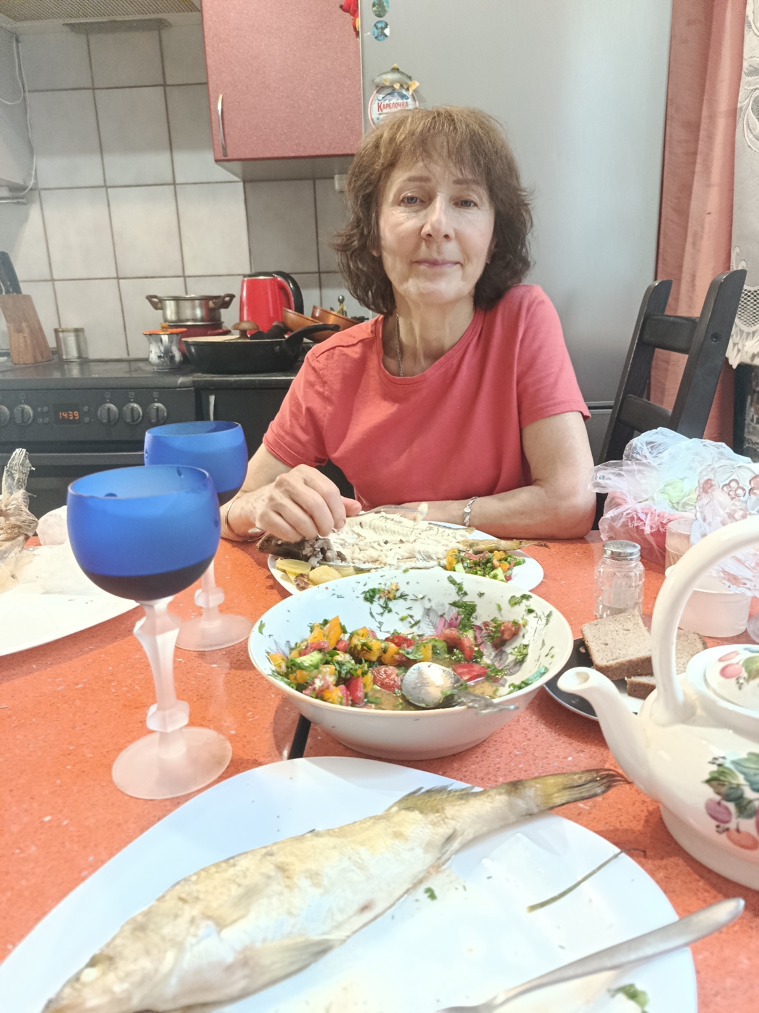 A medium shot shows a woman sitting at an orange table in a kitchen, looking at the camera. She has brown hair and is wearing a coral-colored t-shirt. In front of her is a meal consisting of a whole fish on a white plate, a bowl of chopped salad with tomatoes and herbs, and two blue wine glasses. A teapot and bread are also on the table. The background shows kitchen cabinets, a stove, and a rack with pots and pans. The time on the stove's display reads 14:39. The scene appears to be in Moscow, Russia, based on the presence of Russian text on a label in the background.