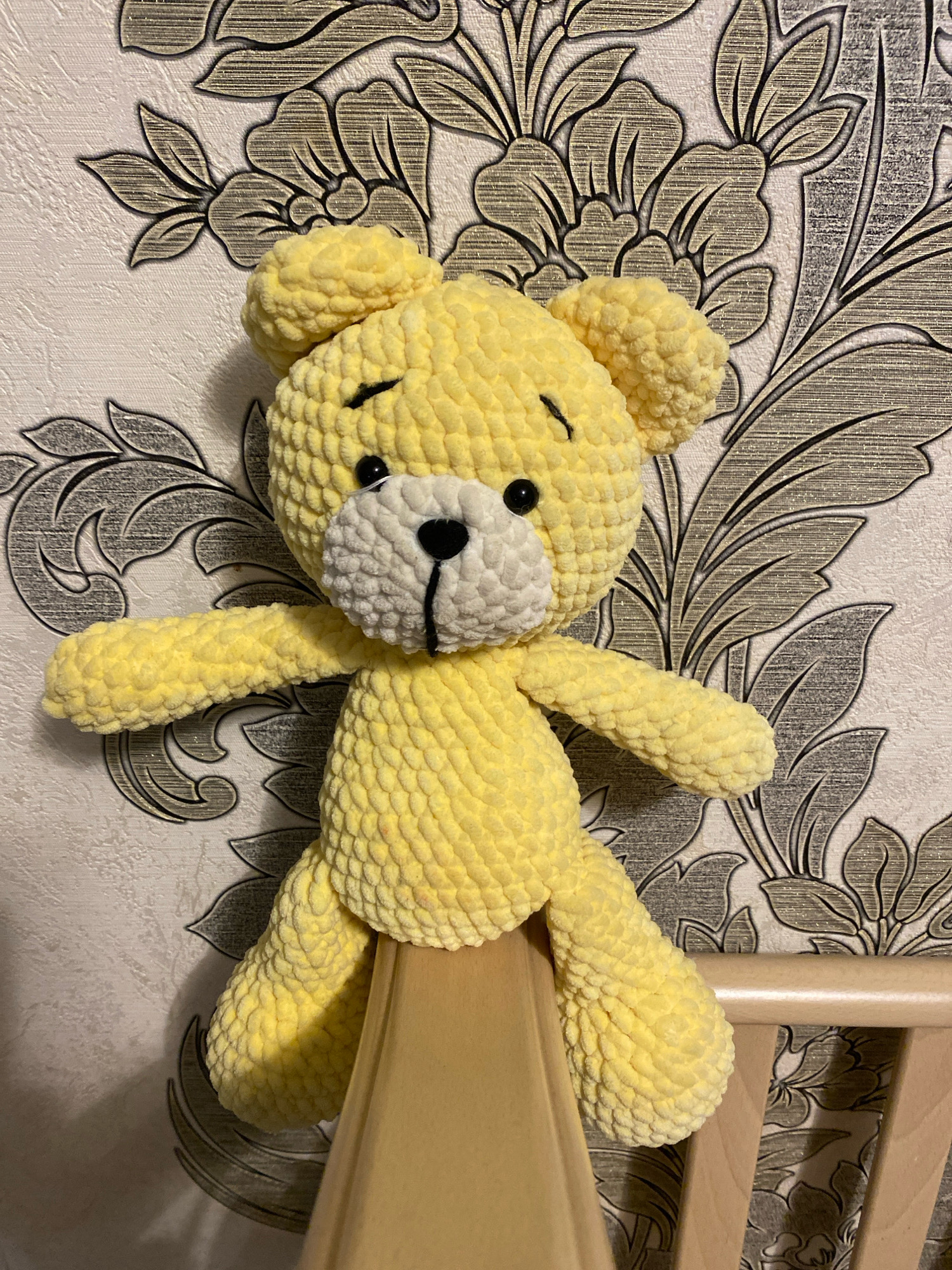 A handmade yellow crocheted teddy bear is positioned on a wooden surface, in front of a wall with a patterned wallpaper. The teddy bear is facing the viewer and has black eyes, a black nose, and a white muzzle. Its arms are outstretched to the sides. The wallpaper behind the bear features a grey floral pattern against a textured white background. The scene is indoors. There is no information to suggest this location is in Korablino, Russia.