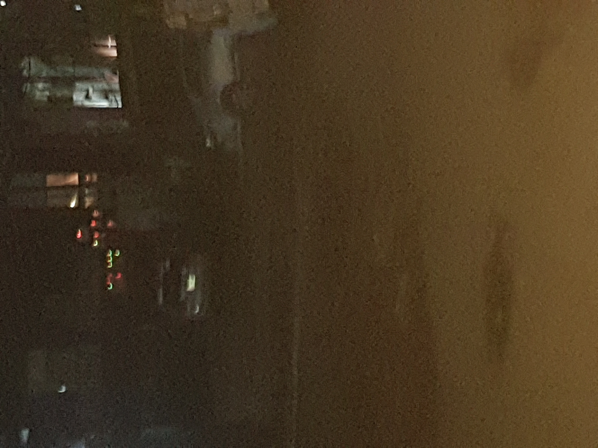 The image shows a blurred view of a city scene at night, likely taken through a window. On the left side of the frame, indistinct buildings and lights can be seen. The right side of the image is largely out of focus, with a muddy brown tone. The red and green lights and blurred objects suggests it could be a view from Shangisha, Nigeria at night. The scene is grainy and dark, with no people visible.