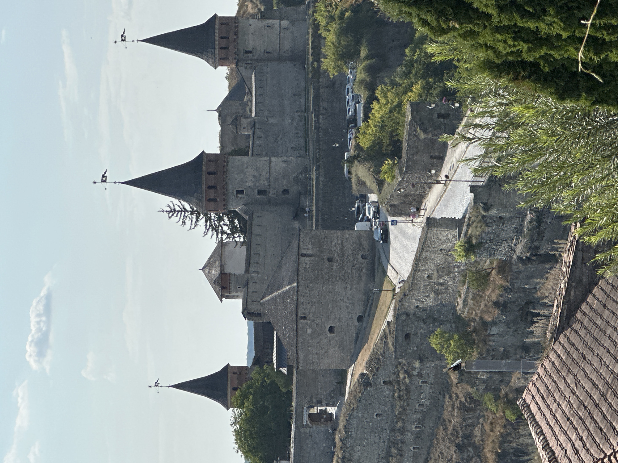 The image shows a panoramic view of the Kamianets-Podilskyi Castle in Kamianets-Podilskyi, Ukraine. The castle is a large, stone fortress with multiple towers, battlements, and a central courtyard. The towers have conical roofs, and some are adorned with weather vanes. The castle is situated on a rocky, elevated terrain, with a road winding its way up towards the entrance. Several cars are parked on the side of the road, and a few people are walking along the path. Green trees and foliage surround the castle, and the sky is partly cloudy. In the foreground, the tiled roof of a building is visible. The overall scene depicts a historic architectural landmark in a natural setting.