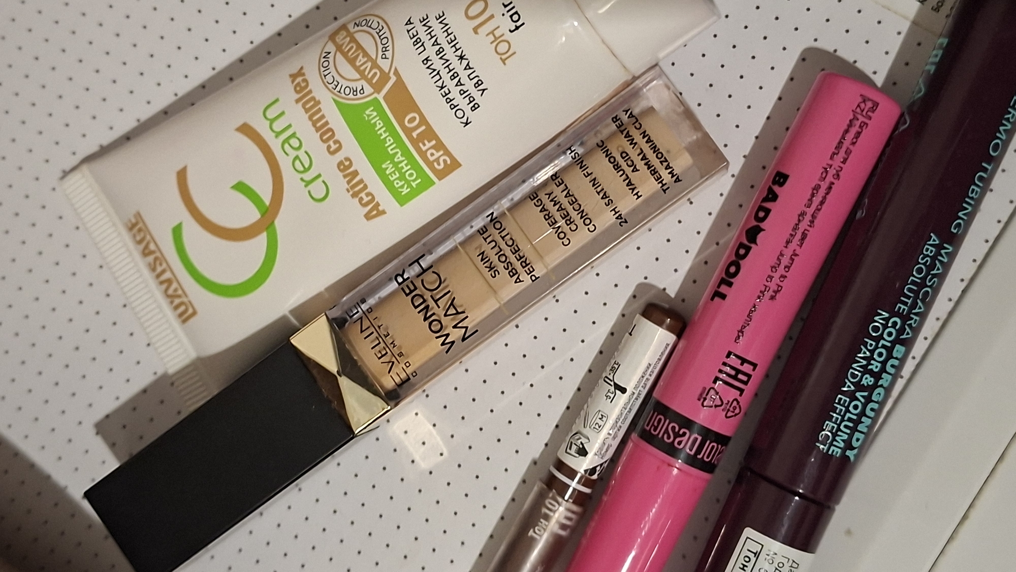 The image displays a collection of cosmetic products laid out on a white surface with a subtle grey polka dot pattern. The products are positioned at an angle, with the top edge of the frame containing a partially visible white tube with text that includes "LUXVISAGE" and "CC cream Active complex". Below this, a clear glass bottle with a black cap and gold accents contains a beige liquid product labeled "EVELINE COSMETICS WONDER MATCH SKIN ABSOLUTE PERFECTION COVERAGE CREAMY CONCEALER 24H SATIN FINISH HYALURONIC ACID THERMAL WATER AMAZONIAN CLAY". To the left of this bottle, a brown cosmetic pencil with white text is visible, some of which reads "ТОН 107". Below the concealer bottle, a bright pink mascara tube is positioned horizontally, featuring branding that appears to be "BADBDLL" and possibly some text in Russian. At the bottom of the frame, a dark purple mascara tube is also laid out horizontally, with white text indicating "ABSOLUTE COLOR & VOLUME NO PANDA EFFECT BURGUNDY". The overall scene is a still life arrangement of these beauty products. There is no indication of the location being in Забалоцкі сельскі Савет, Belarus or any other specific geographical location.