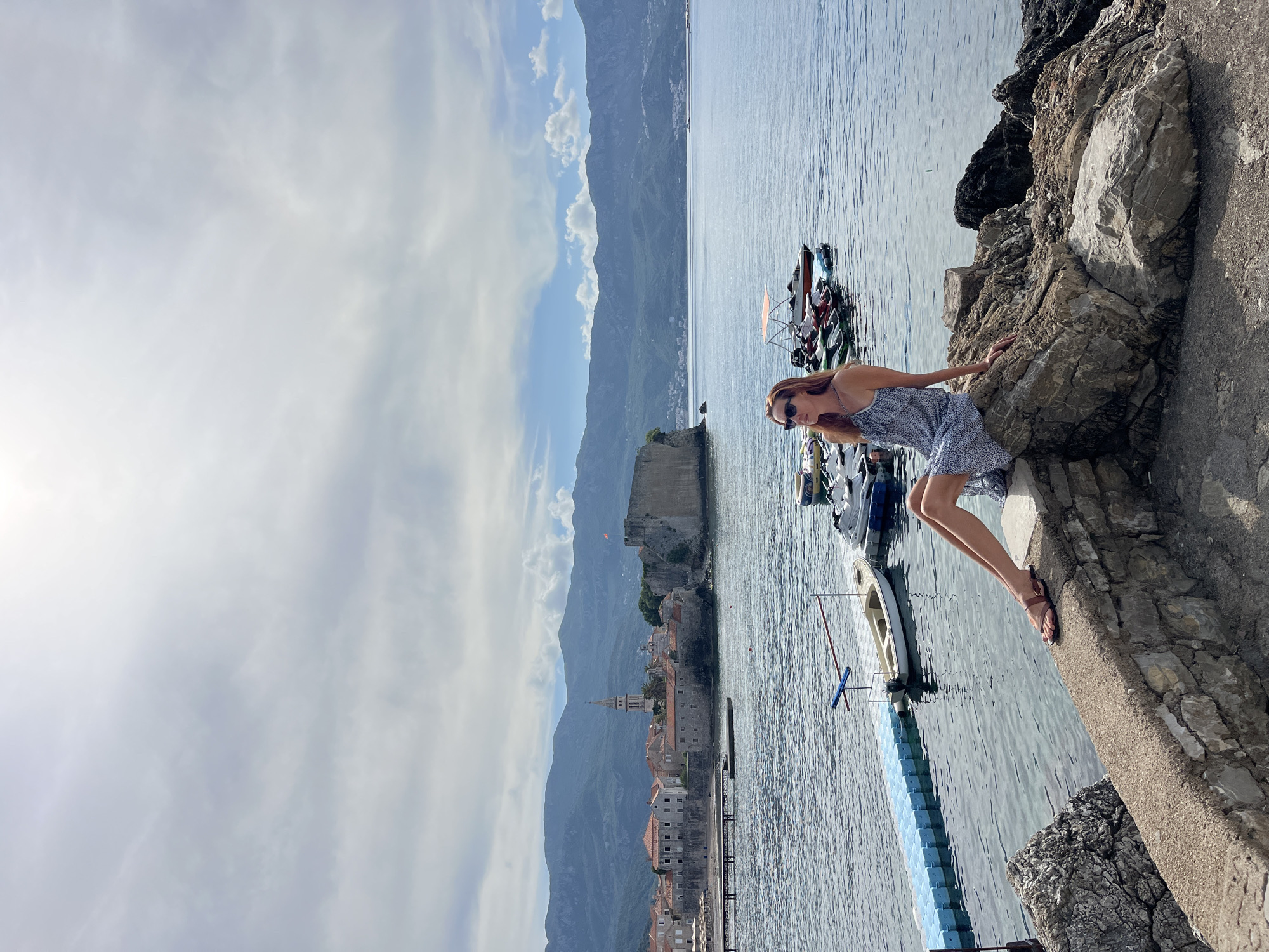 The image, correctly oriented, depicts a serene coastal scene in Budva, Montenegro, on a partially cloudy day. In the foreground, a woman with long, reddish-brown hair, sunglasses, and a patterned sundress is posing. She leans against a textured rock formation on the shoreline, her left hand resting on the rock, and looks towards the camera with a gentle expression. Her legs extend towards the calm, blue-grey water, and she wears sandals.
To her right, the water is populated with several boats, including motorboats and jet skis, moored alongside a light blue floating dock. Beyond the immediate waterfront, the historic architecture of Budva Old Town is visible, featuring numerous buildings with terracotta roofs, a prominent bell tower with a spire, and an ancient fortress wall. This picturesque town is nestled at the base of towering, lush mountains that stretch across the horizon under a sky dotted with white clouds. The scene conveys a sense of peaceful coastal beauty. No visible text is present in the image.
