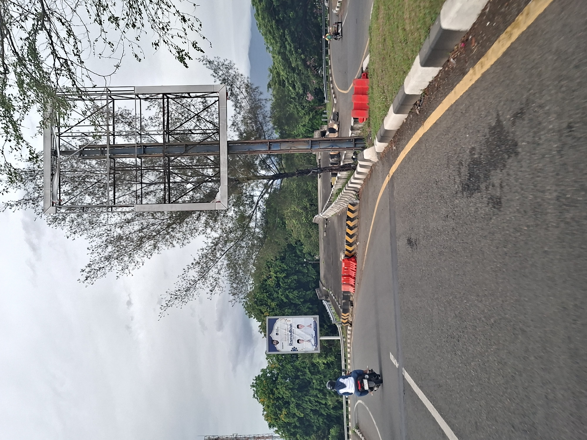 The image captures an overcast day on a curved road in Tanjong, Indonesia. A motorbike with a rider wearing a white and blue jacket and helmet is moving on the road, with a double white line separating traffic. A billboard displaying an advertisement for "Ramadhan" stands beside the road, against a backdrop of lush green trees and a cloudy sky. An unfinished billboard frame is visible in the upper-left corner. The road is lined with red and white traffic markers, and black and white patterned curbs. Beyond the road, the scene transitions into dense foliage leading up to a distant, hazy mountain range.
