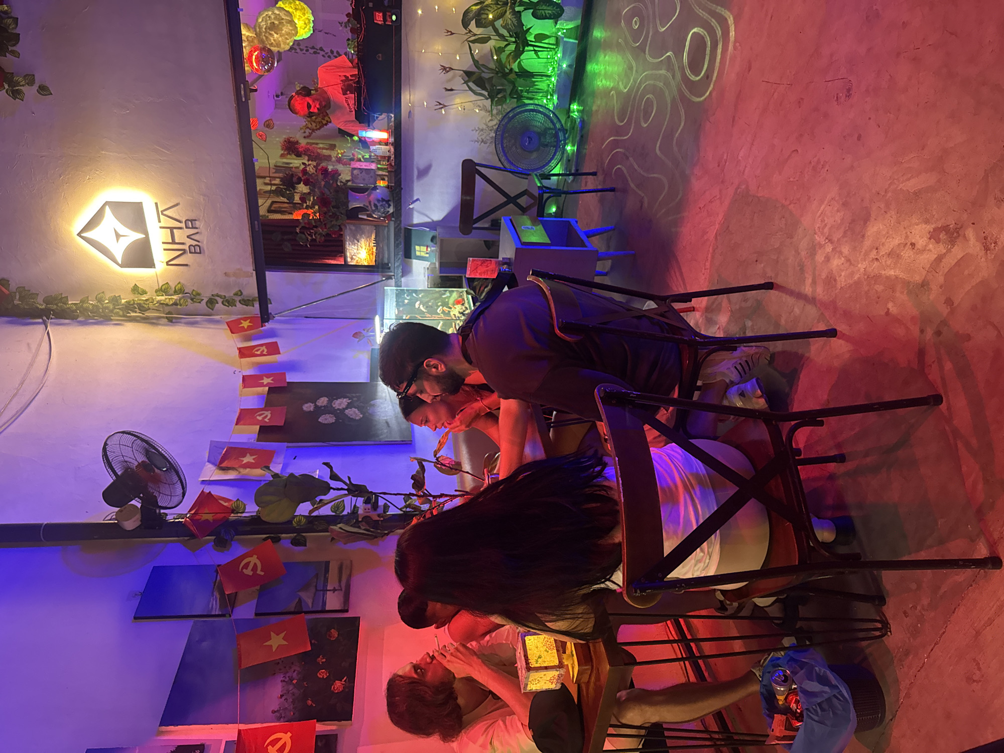 This is an indoor evening or night scene depicting a vibrant bar or lounge, likely in Đà Nẵng, Vietnam. The space is bathed in dynamic artificial lighting, casting strong red, purple, and green hues across the concrete floor and walls. Swirling patterns are projected onto the floor, enhancing the energetic ambiance.
Several individuals are present, suggesting a social gathering. Towards the back, a DJ wearing headphones is visible behind a console, indicating that music is being played. In the foreground and mid-ground, a group of approximately five patrons are seated at small tables, facing each other and socializing. One person with long dark hair is seen from behind, while others are partially visible from the side or front, engaged in conversation. One individual appears to be smoking.
The bar features a glowing, stylized diamond-shaped sign on a light wall, with the text "NHA BAR" written vertically beside it. Artificial greenery is draped below the sign. Along another wall, miniature flags are strung up as decorations; these include the red flag with a yellow star (flag of Vietnam) and red flags adorned with the hammer and sickle (symbol of the Communist Party of Vietnam). Other objects include plants, electric fans, chairs, tables, and what appears to be a trash bin. The setting suggests a casual and lively atmosphere.
