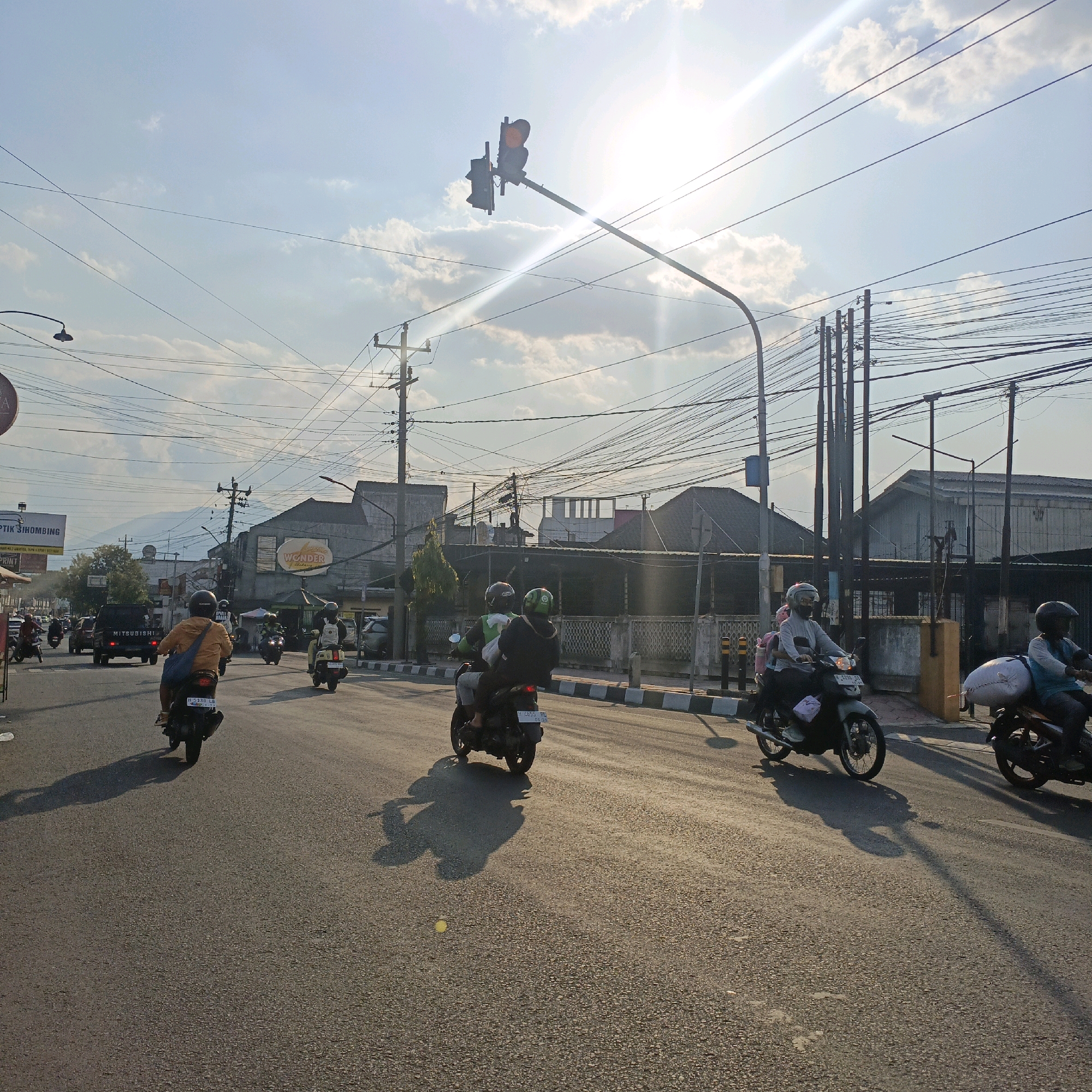 This image depicts a street scene in Salatiga, Indonesia, characterized by its urban infrastructure and daily life activities. The sun is low in the sky, casting long shadows and creating a warm, bright atmosphere with lens flare evident. Several motorcycles are in motion on the asphalt road, some carrying one or two passengers. The people on the motorcycles are wearing helmets for safety.
The road is lined with buildings, some appearing to be commercial establishments with signage, including one that reads "WONDER Bakery". Utility poles with a dense network of wires crisscross the sky, a common sight in Indonesian cities. A traffic light is visible, positioned on a tall pole. In the background, mountains are faintly visible, suggesting the city's geographical setting. The sidewalks have a checkered pattern, and a few trees are present. The overall impression is of a busy street in a city during daylight hours, with the sun adding a dramatic effect to the scene.