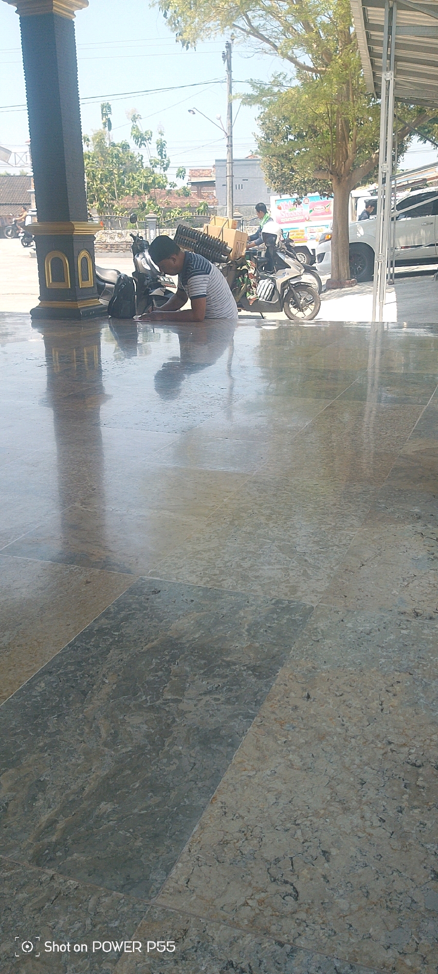In the city of Weru, Indonesia, a young man with dark hair, wearing a striped shirt and a black cap, is sitting on a polished floor, hunched over and writing in a notebook. He is positioned to the left of two motorcycles, one with a load of boxed items secured on its rear. A backpack lies on the ground near him. In the background, another person is on a motorcycle, and beyond them, a white van is parked. The scene appears to be in an open area with a covered structure overhead and trees and buildings visible in the distance. The floor is made of large, reflective tiles with a mottled pattern, creating significant reflections of the overhead structure and the people present.