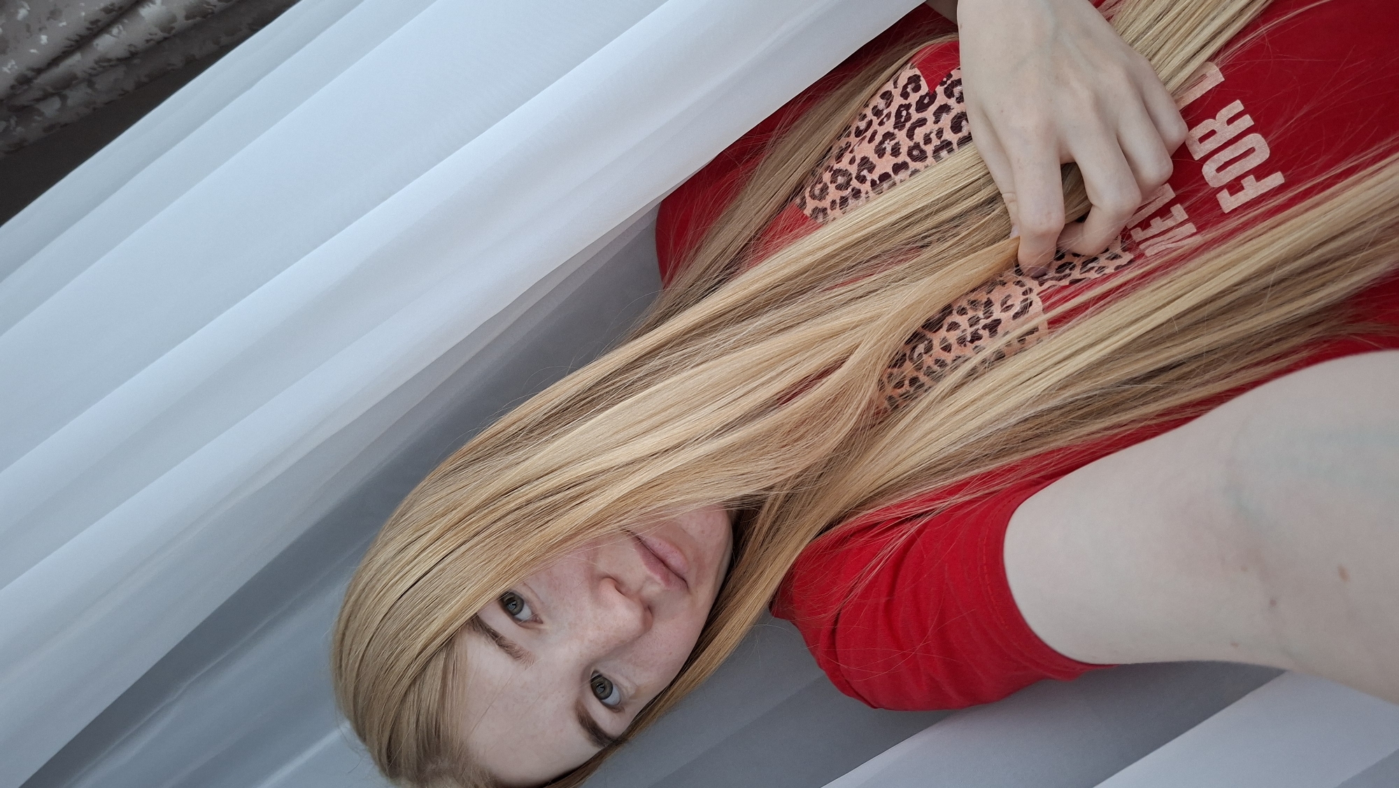 A woman with long, blonde hair is lying down, looking at the camera. She is wearing a red t-shirt with an animal print design and white lettering. Her hand rests on her chest, entangled in her hair. The background consists of white, sheer curtains. The location is Chaikovsky, Russia.