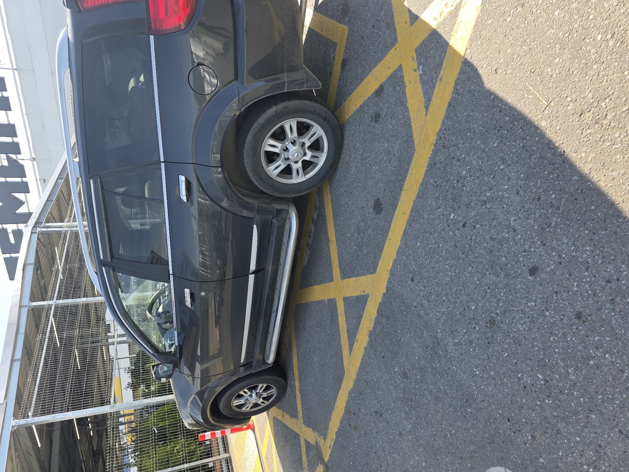 A dark gray SUV is parked over a yellow crisscross marking on asphalt. The vehicle is positioned at an angle, with its front wheels turned slightly. The parking lot appears to be outdoors, next to a building with a metal structure and a wire fence. There is a red and white traffic cone near the front of the vehicle. Sunlight casts a shadow from the car onto the ground.