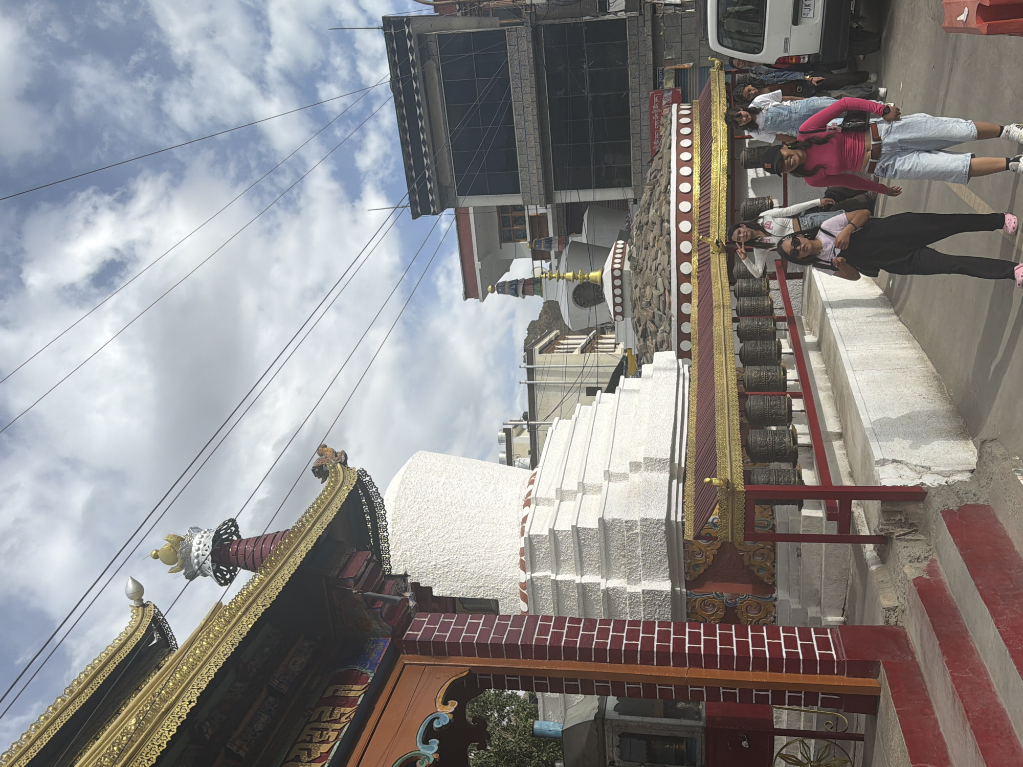 In Leh, India, a group of young women walk along a sidewalk next to a Buddhist stupa adorned with prayer wheels. A white stupa with a tiered base is visible on the left, with a colorful entryway featuring red brick accents and golden decorative elements. Several women are walking towards the viewer, some looking at the camera. One woman in the foreground wears a pink long-sleeved shirt and denim shorts, while another to her left wears a black jumpsuit. Behind them, more women are walking, one in denim overalls. The background shows modern buildings and a clear sky with scattered clouds. A white vehicle is parked on the right side of the road.