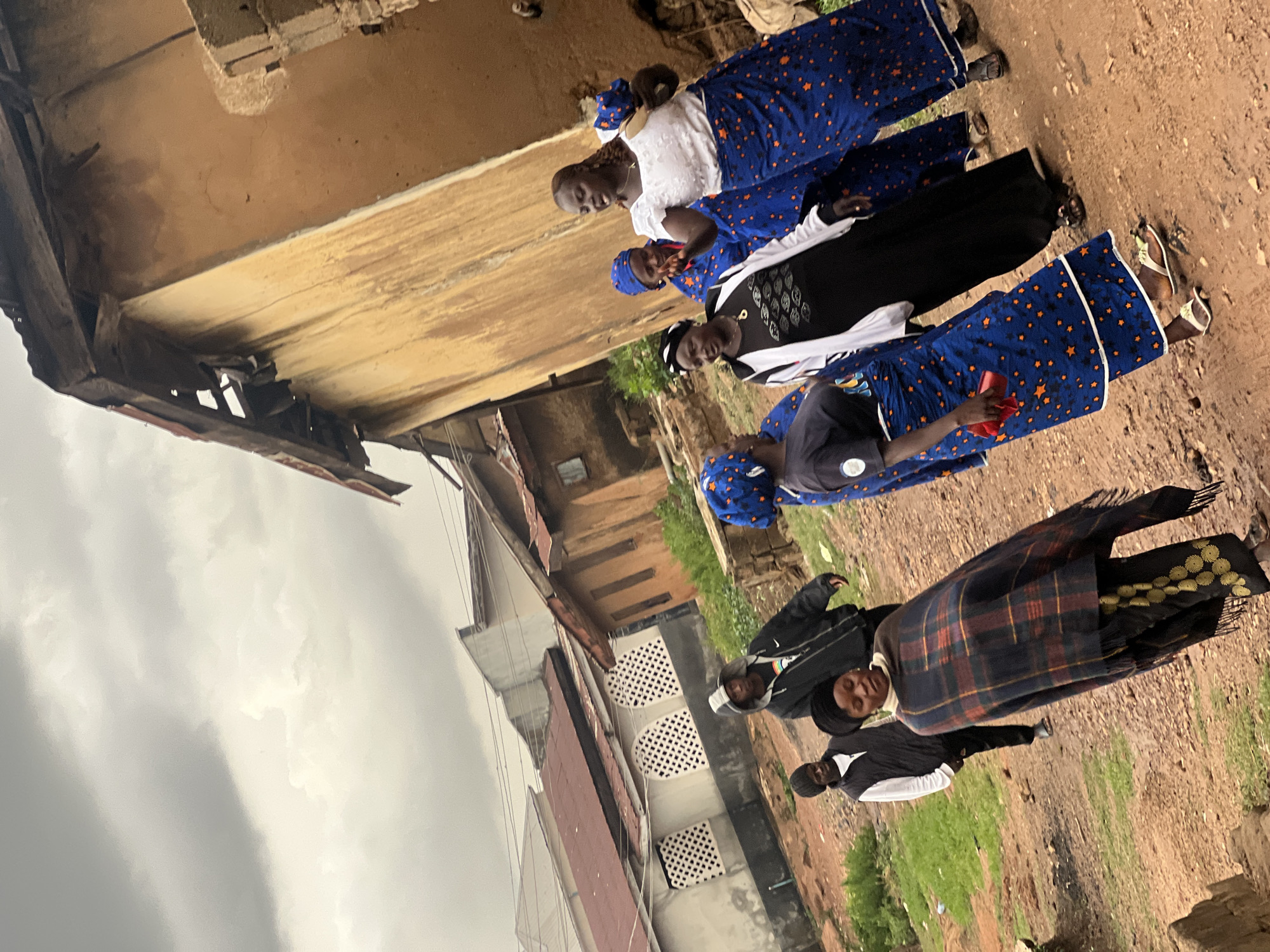 The image depicts a group of individuals in what appears to be Gura Top, Nigeria. In the foreground, four women stand shoulder to shoulder, wearing matching blue skirts with gold star patterns, and headscarves. One woman is wearing a white top, and another is holding a red object. Behind them are two men. One of the men is draped with a tartan plaid blanket over his shoulders, wearing a wide-brimmed hat. The other man is wearing a black and white track suit and a black beanie.
The backdrop shows an urban environment, including aged, weathered buildings, some constructed with exposed brick and stucco. An elevated structure with damaged eaves is also visible in the upper portion of the frame. The ground appears to be a mixture of soil and gravel. Overcast skies suggest it might be early morning or late afternoon, or the weather may be cloudy.