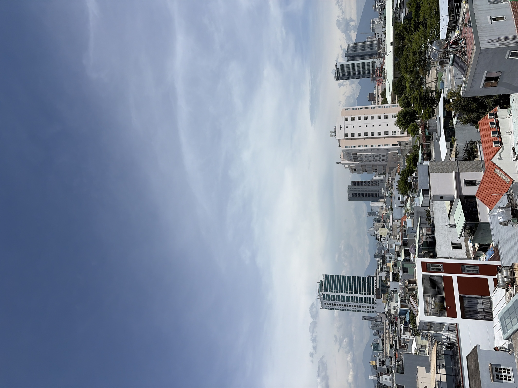 A daytime, high-angle, eye-level shot captures a sprawling cityscape under a bright, partly cloudy sky. The foreground is dominated by the rooftops of numerous buildings, featuring a mix of architectural styles and materials, including white, grey, and red-tiled roofs. Some rooftops are equipped with water tanks and solar panels. Several buildings have prominent balconies and window structures.
Midground of the image showcases a dense urban environment with a variety of residential and commercial buildings of varying heights. Prominent among these are several taller, modern buildings, including a distinctive white and green striped skyscraper to the left, and a pale pink rectangular skyscraper with numerous windows to the right of center. These taller structures suggest a more developed area within the city.
The background reveals a distant mountain range, partially obscured by haze and clouds, adding depth to the scene. A few cranes are visible, indicating ongoing construction within the city. The overall impression is of a vibrant, developing coastal city. The image is consistent with a view of Đà Nẵng, Vietnam, given the presence of modern skyscrapers, dense urban development, and the proximity of mountains to the coast. No people are visible in the frame.