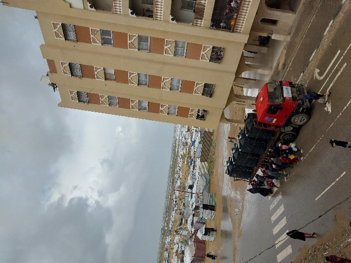 An elevated view captures a street scene in Khan Yunis, Palestinian Territory, under a heavily overcast sky. A red flatbed truck, featuring a white cab and an apparent UNICEF banner on its front bumper and an EU logo on its side, is parked on a wet asphalt road. Its bed is loaded with approximately two dozen dark, possibly black or dark blue, plastic water tanks arranged in multiple stacks.
Approximately 15-20 individuals, including adults and children, are clustered around the left side of the truck, engaged in filling various personal containers such as buckets, jerrycans, and bottles with water, suggesting a water distribution operation. The road surface exhibits puddles and reflections from recent rain, with clear white pedestrian crosswalk stripes and a curved arrow road marking visible. To the right, a multi-story tan residential building with patterned window surrounds, balconies, and a recessed ground floor supported by arches rises prominently. A white water tank is visible on the building's roof. To the left of the road and extending into the distant background, a sprawling informal settlement or displacement camp is visible, characterized by numerous white and grey tents and makeshift shelters spread across muddy, uneven terrain. Several individuals are seen walking on the wet ground near the camp.