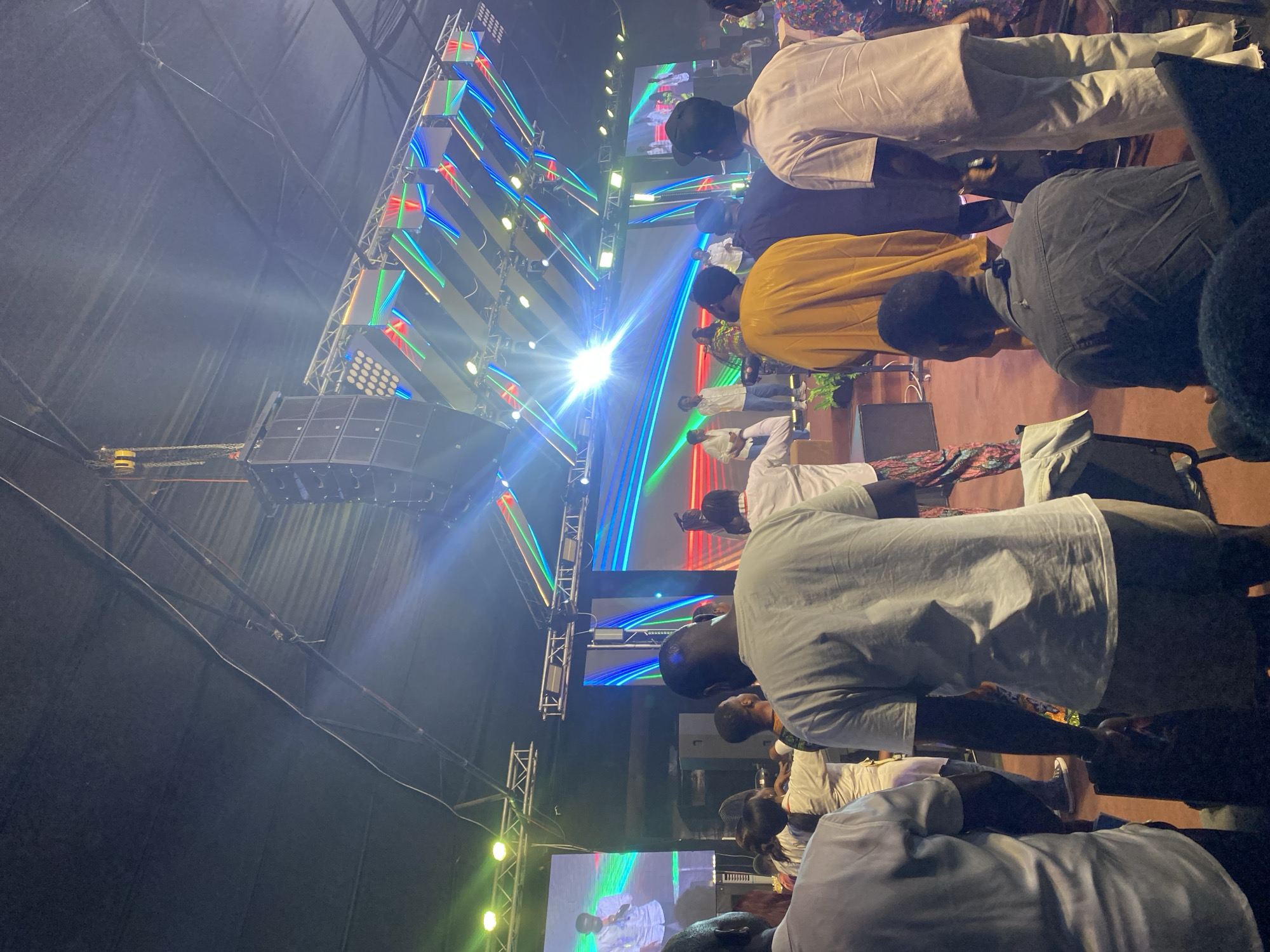 The image captures an indoor event, likely a concert or religious gathering, held within the city of Ado, Nigeria. The scene is dominated by a brightly lit stage featuring a large screen displaying an image of a man in a white shirt holding a microphone, and an array of colorful, illuminated geometric structures suspended from the ceiling, with prominent speakers mounted on the left side. A crowd of people, viewed from behind, fills the foreground, their attention directed towards the stage. The individuals are dressed in casual attire, with some wearing white shirts, and the environment appears to be a large hall or auditorium with a dark ceiling and a red carpeted floor. The lighting is dramatic, with bright stage lights creating lens flares and illuminating the scene.