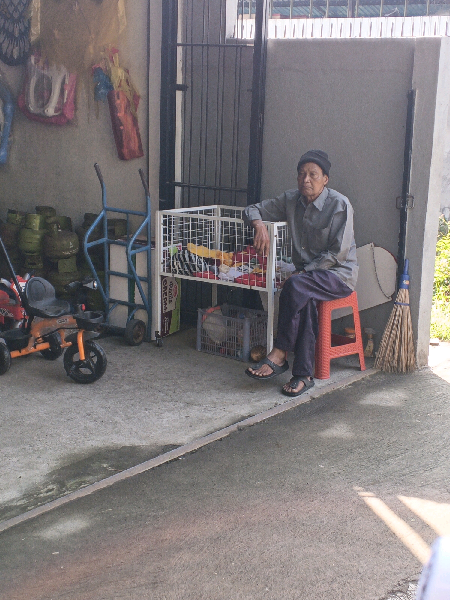 A Balinese man wearing a dark beanie and a gray long-sleeved shirt sits on a red plastic stool in front of a white metal shelf unit. The man is positioned in what appears to be an outdoor area, possibly a driveway or a loading dock, adjacent to a building with a textured gray wall and a dark metal gate. He is leaning his left arm on the shelf unit. The shelf unit contains various items, including what appear to be fabrics and boxes. To the left of the shelf unit are several green gas cylinders stacked and a blue hand truck. In the foreground, a small orange and black child's tricycle is visible. To the right of the man, a broom leans against the wall. The ground is a concrete surface, with some areas appearing wet or stained. The overall scene suggests a utilitarian or storage space within a residential or commercial area in Penebel, Indonesia.