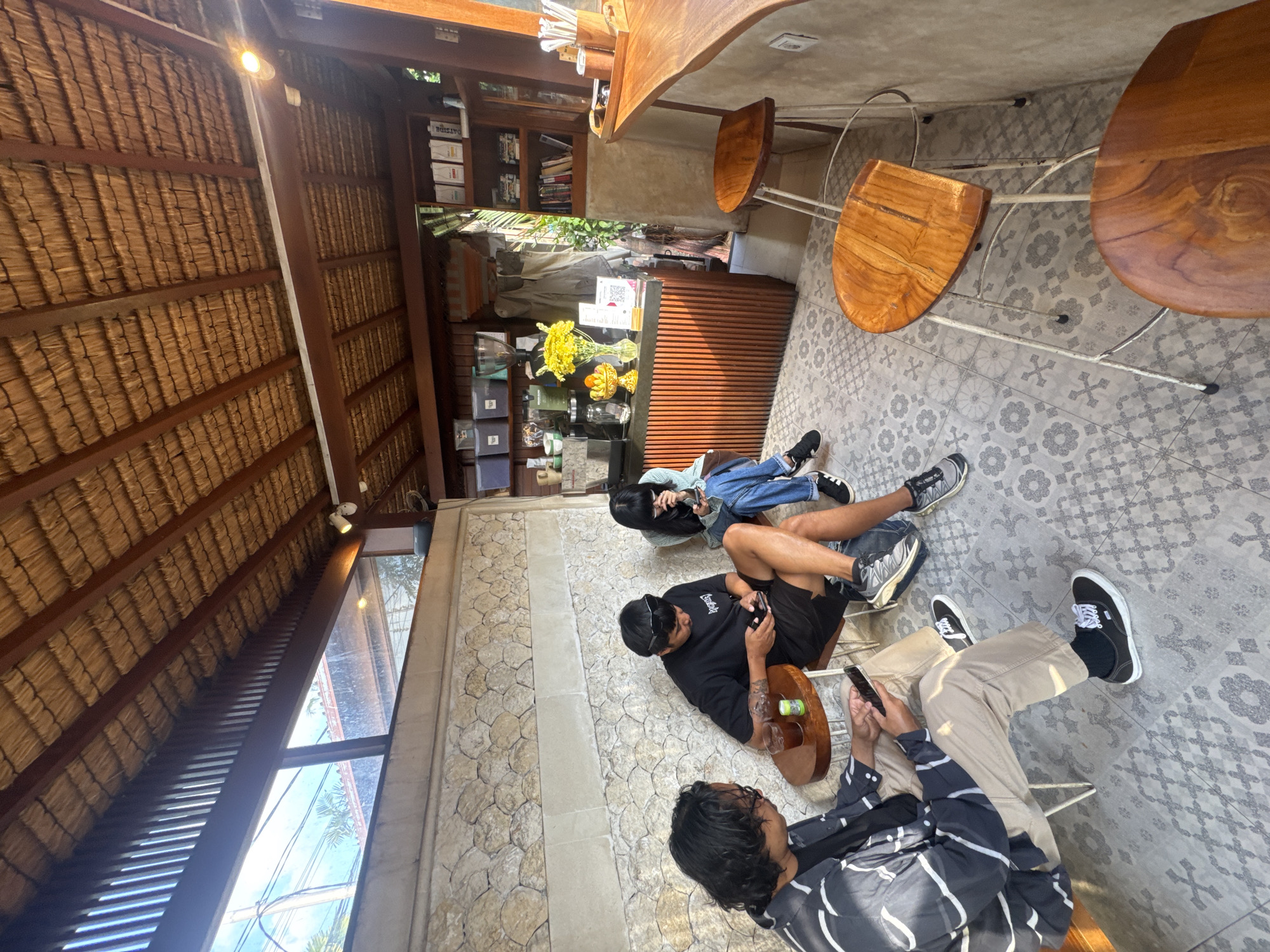 This image depicts the interior of a cafe in North Kuta, Indonesia. Three individuals are seated, engaged with their mobile devices. One person on the left is wearing a striped jacket and khaki pants, holding a smartphone. The person in the center, wearing a black t-shirt and black shorts, is also looking at their phone. The third person on the right, wearing a blue long-sleeved shirt and blue jeans, is also interacting with their phone.
The cafe features a thatched roof supported by wooden beams, and large windows that allow natural light to filter in. The walls are a textured stone material. In the background, a counter is visible with coffee-making equipment, shelves stocked with books and various items, and a vase of yellow flowers. The seating includes small round tables and wooden stools with metal legs. The floor is tiled with a patterned design. The overall atmosphere suggests a casual and relaxed environment.
