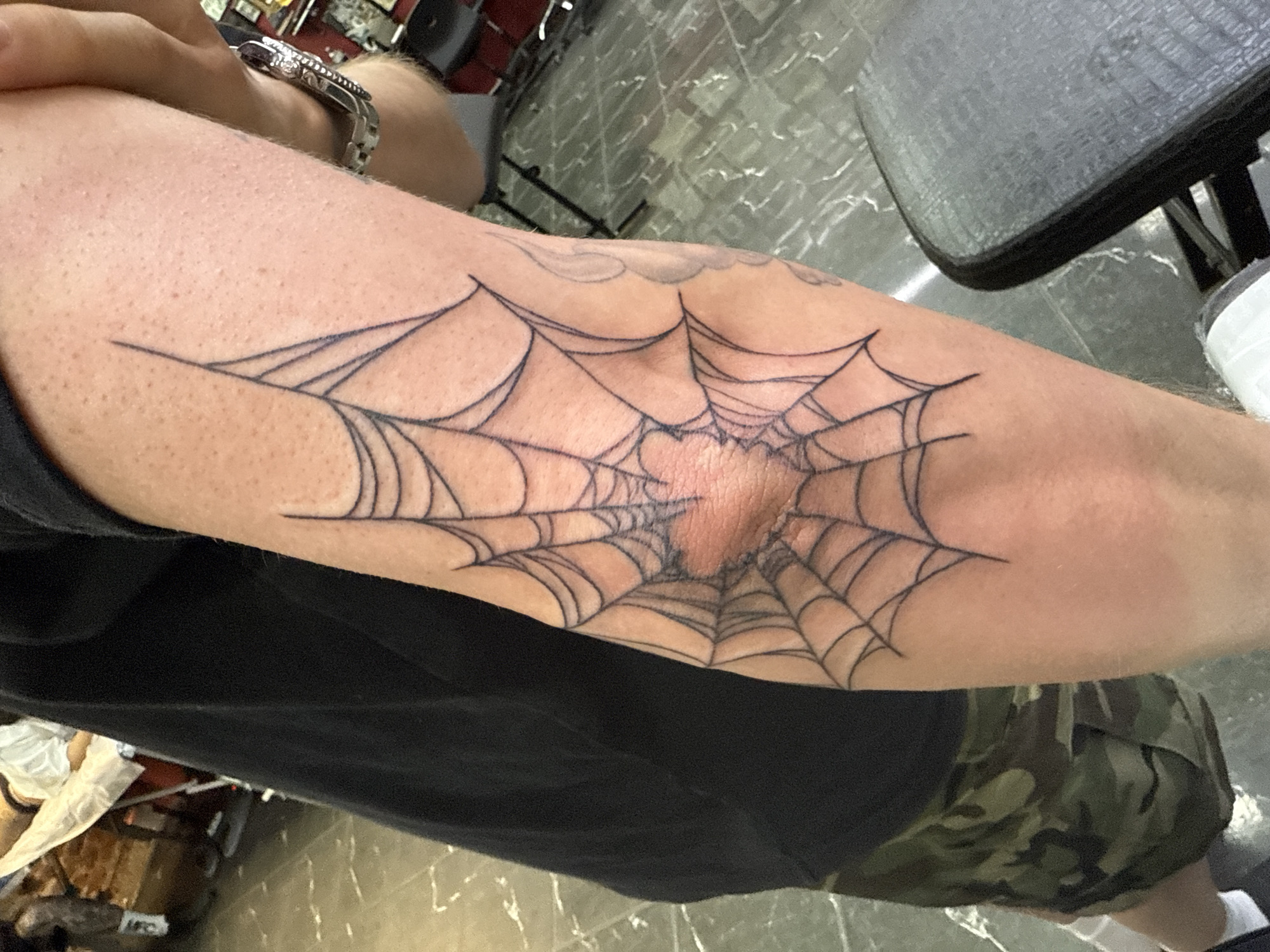 A close-up shot captures a person's arm, featuring a spiderweb tattoo centered around the elbow. The tattoo is executed in black ink, with the elbow joint forming the center of the web. Above the web, a faint outline of a cloud tattoo is visible. The person is wearing a silver wristwatch on the left arm. The individual is also wearing a black t-shirt and camouflage shorts. A black chair with a gray seat is partially visible in the background. The floor beneath the chair appears to be a light-colored tile. The location is indeterminate.