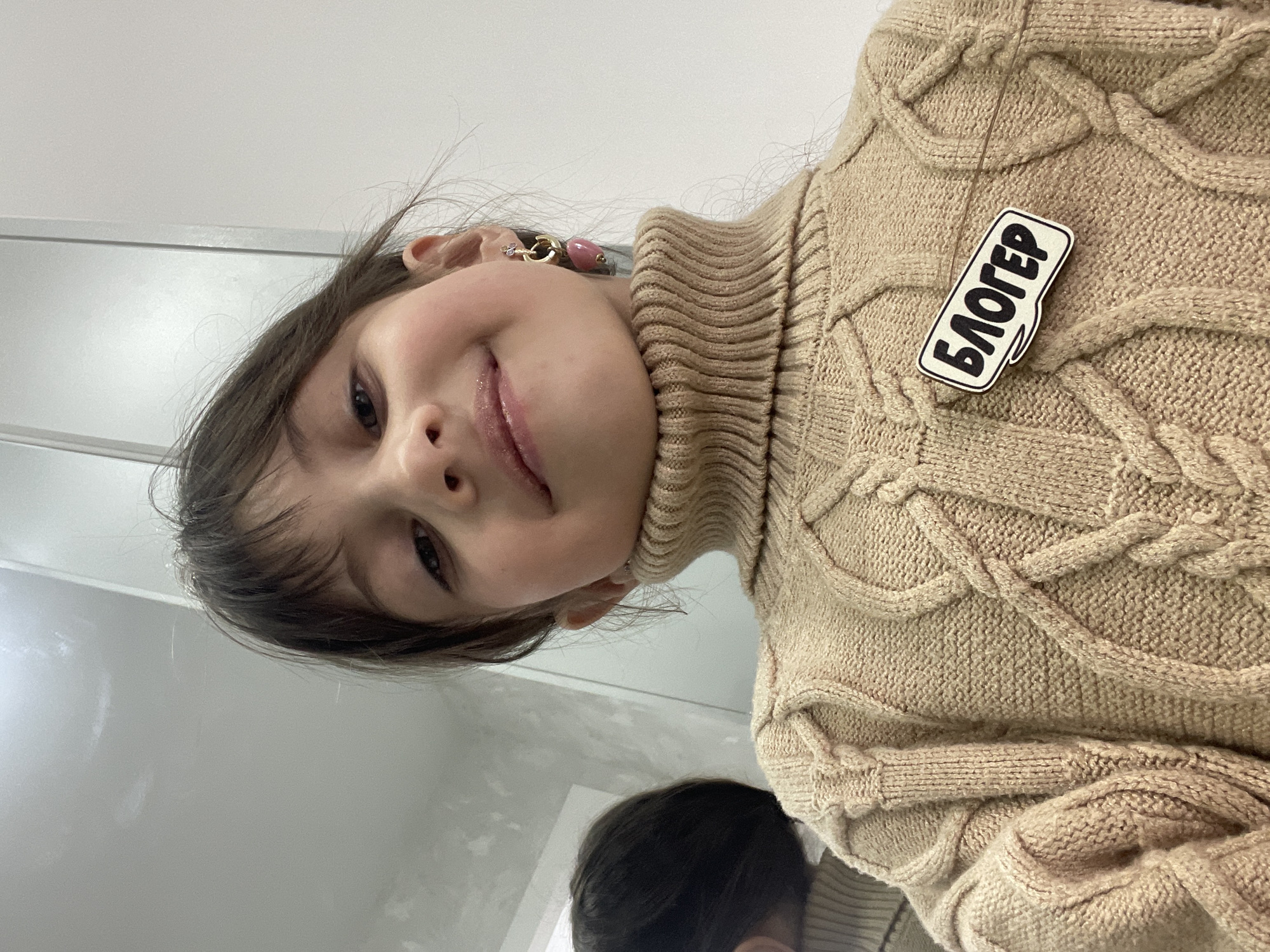 A young girl is taking a selfie from a high angle. She has light skin, dark hair, and is wearing a beige cable-knit turtleneck sweater. A badge with the Russian word "Блогер" (Blogger) is pinned to her sweater. She's wearing a pink earring. The background is a blurred white surface, possibly a wall or ceiling. A partial reflection of another person is visible in the lower part of the frame. There is no information that suggests this is Izhevsk, Russia.