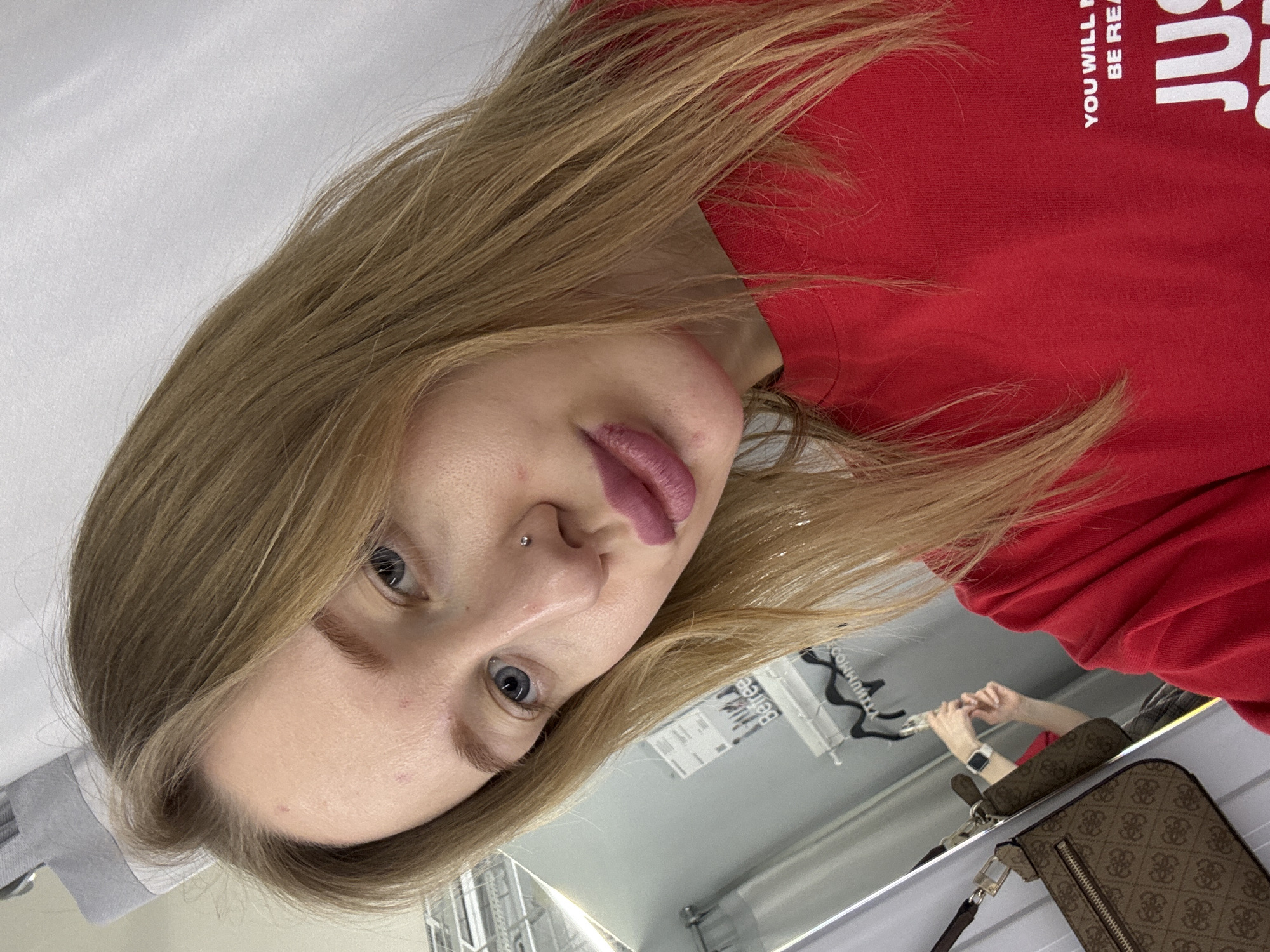 A young, fair-skinned woman with blonde hair is looking at the camera. She is wearing a red sweatshirt with white text printed on it. She has a nose piercing and is wearing lipstick. The background appears to be a room with a mirror reflecting the woman holding her phone, and a handbag. Based on object detection, the handbag has a design suggestive of the "Guess" brand. The location is suggested to be Rostov-on-Don, Russia.