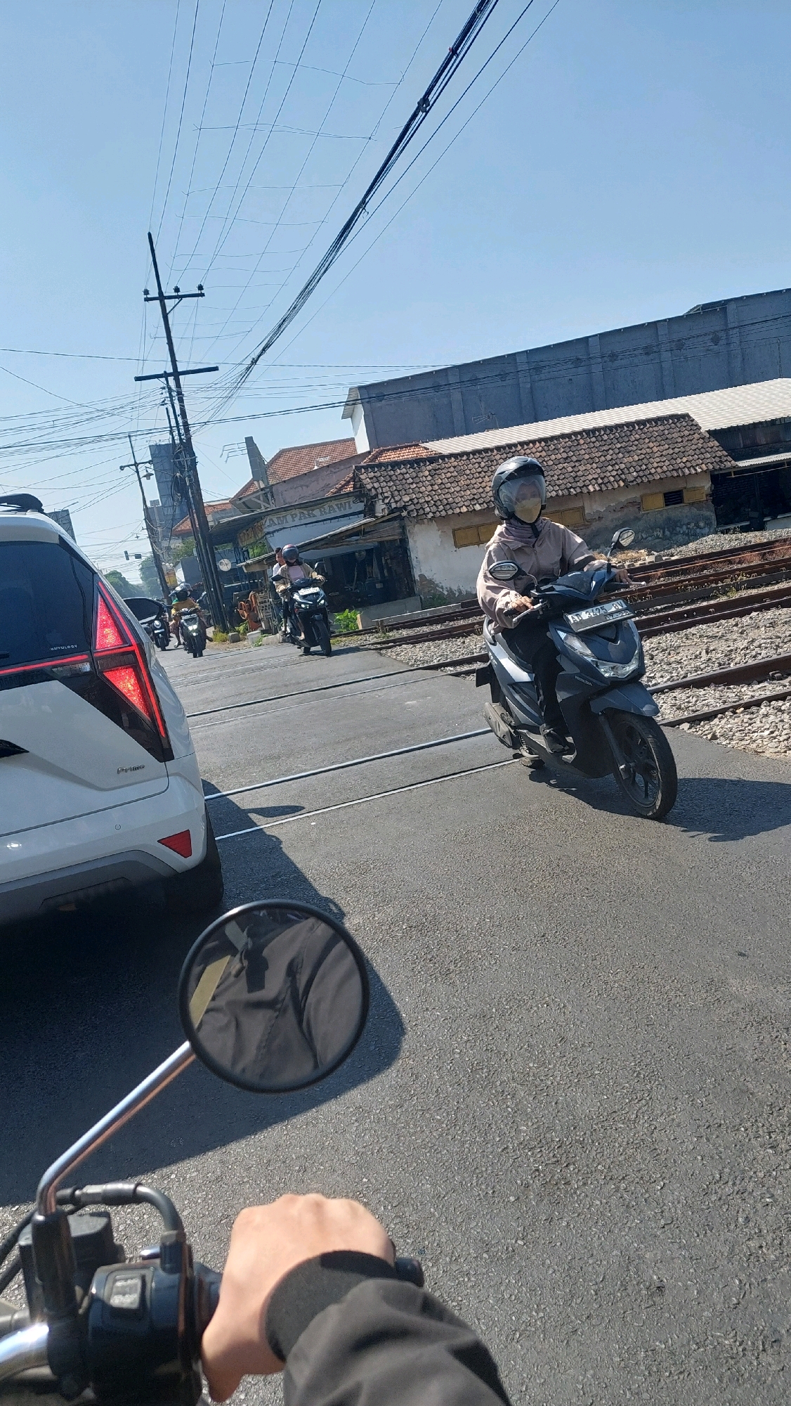 The image captures a sunny day scene at a railway crossing in Sidoarjo, Indonesia, likely on Jalan Raya Porong. In the foreground, the perspective is from a motorcycle, with a hand gripping the handlebar and a side-view mirror reflecting a dark jacket. A white SUV is parked to the left, its taillight visible. Beyond the SUV, several individuals are riding motorcycles, navigating the crossing. One motorcyclist in the mid-ground wears a light brown jacket and a helmet with a yellow mask, riding a dark scooter with license plate "AT 3175 AW". Behind them, other motorcyclists are also visible. The railway tracks run horizontally across the scene, with gravel and ties visible. Buildings with tiled roofs and corrugated metal structures line the background. Overhead, a dense network of electrical wires is strung between poles, characteristic of an urban or semi-urban Indonesian environment. The sky is clear and blue.