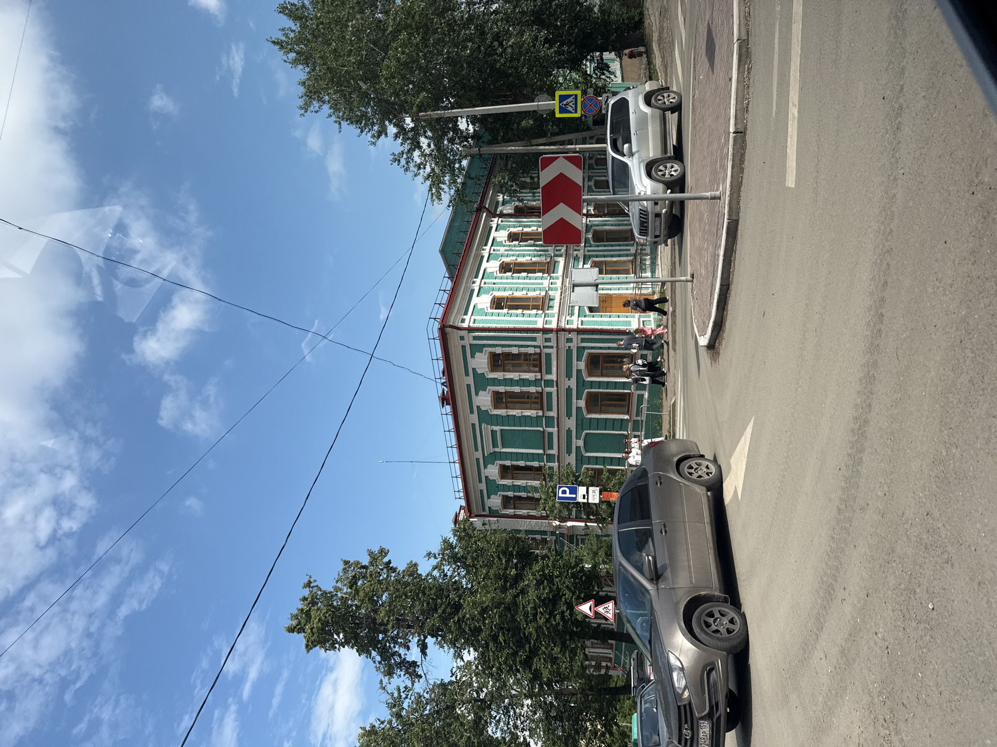 The image shows a street scene in the city of Perm, Russia. The primary subject is a green, two-story building with white decorative trim and brown-framed windows. In front of the building, there is a traffic intersection with several cars and pedestrians. A silver SUV is parked to the right of the intersection, and a gray sedan is driving through it. People are walking on the sidewalk and crossing the street. Traffic signs, including a parking sign and a directional arrow sign, are visible. Trees line the street, and a blue sky with scattered clouds is overhead. The overall impression is a typical urban environment on a sunny day.